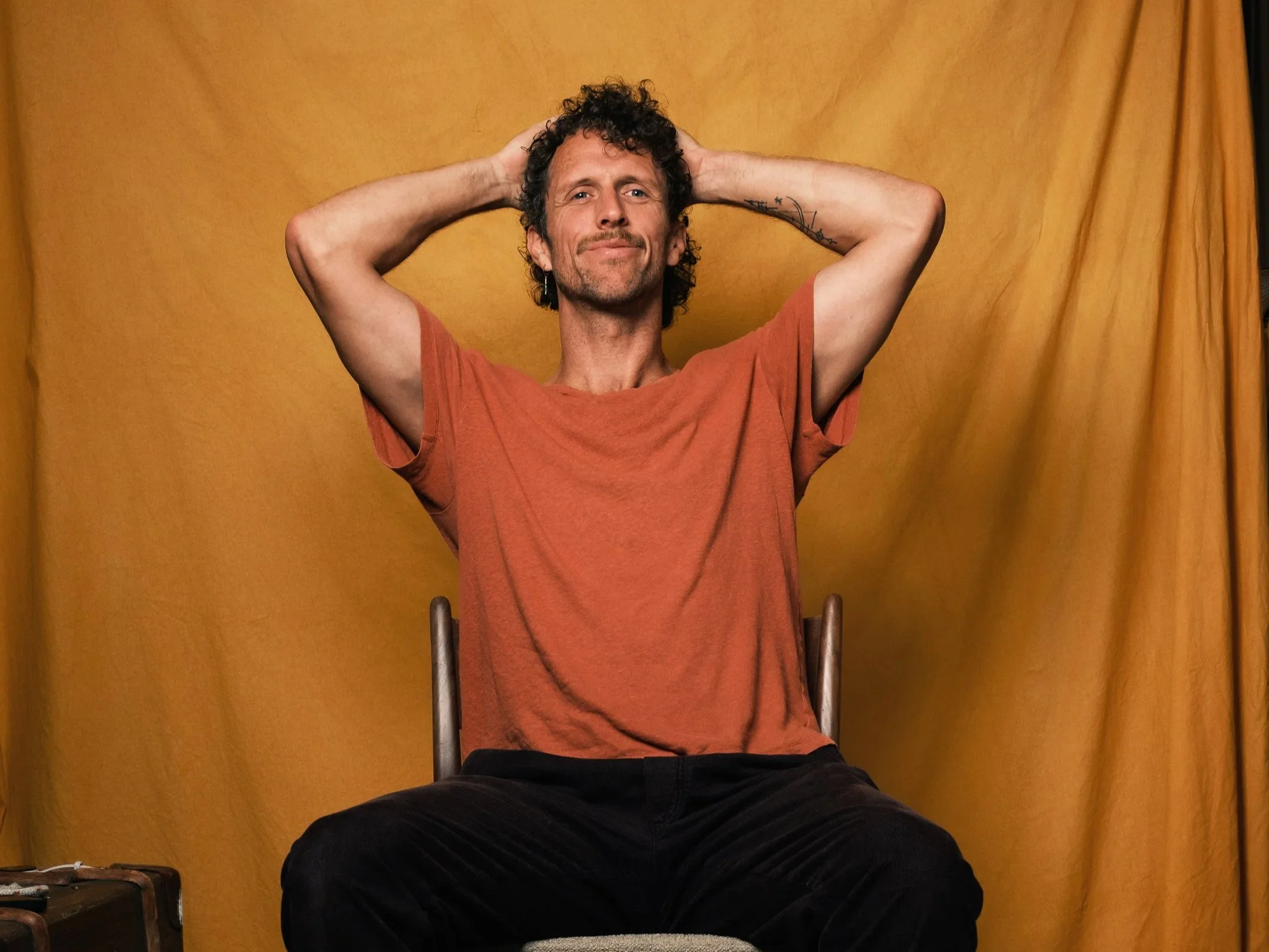 Man met krullend haar zit op een stoel, relaxte pose met handen achter hoofd, oranje t-shirt, gele achtergrond.