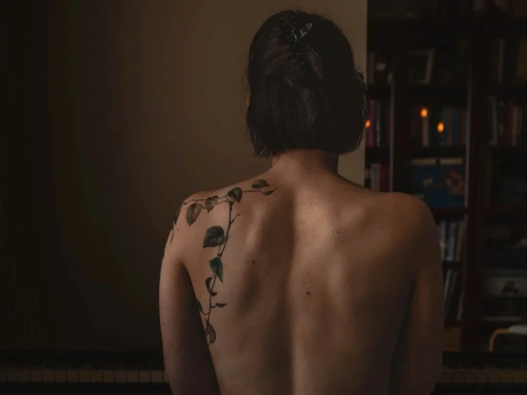 Een vrouw gezien vanaf de achterkant, zonder kleding, met een tattoo van een klimopblad op haar linkerschouder en rug, zittend voor een boekenkast.