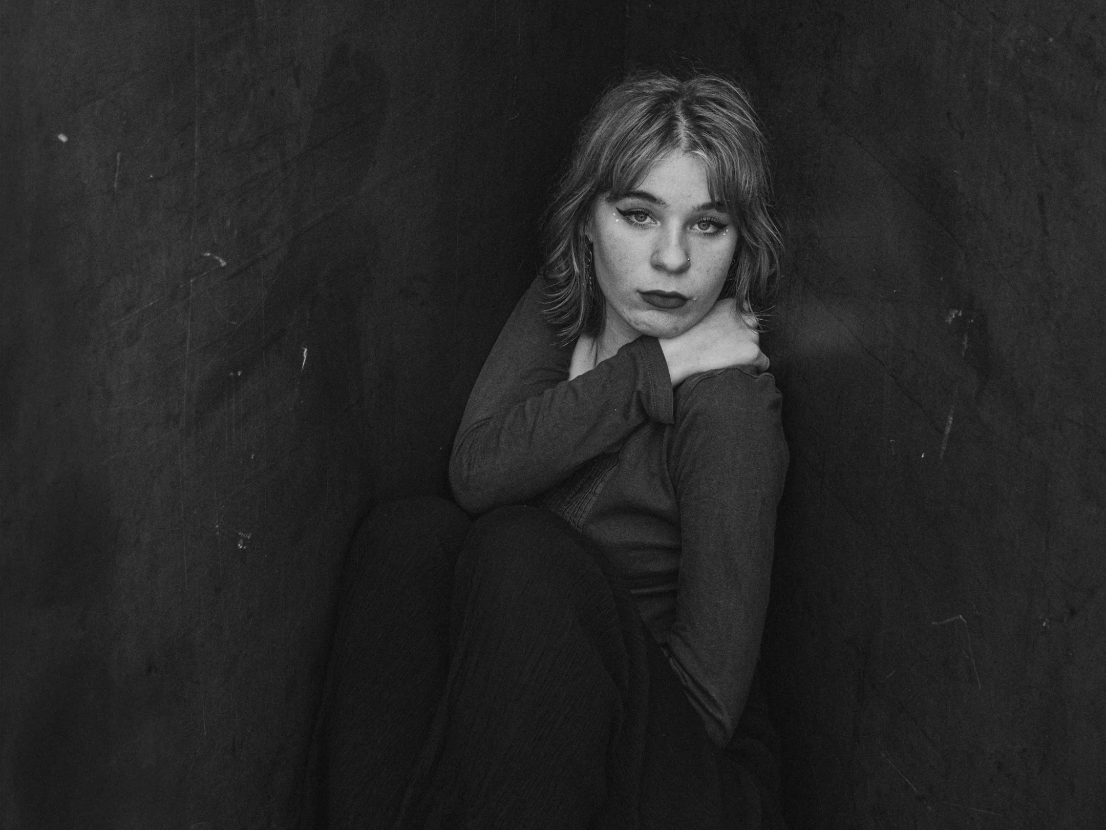 Portret van een jonge vrouw met lichte huid, halflang haar en donkere lipstick, zittend tegen een donkere muur, met een serieuze uitdrukking.