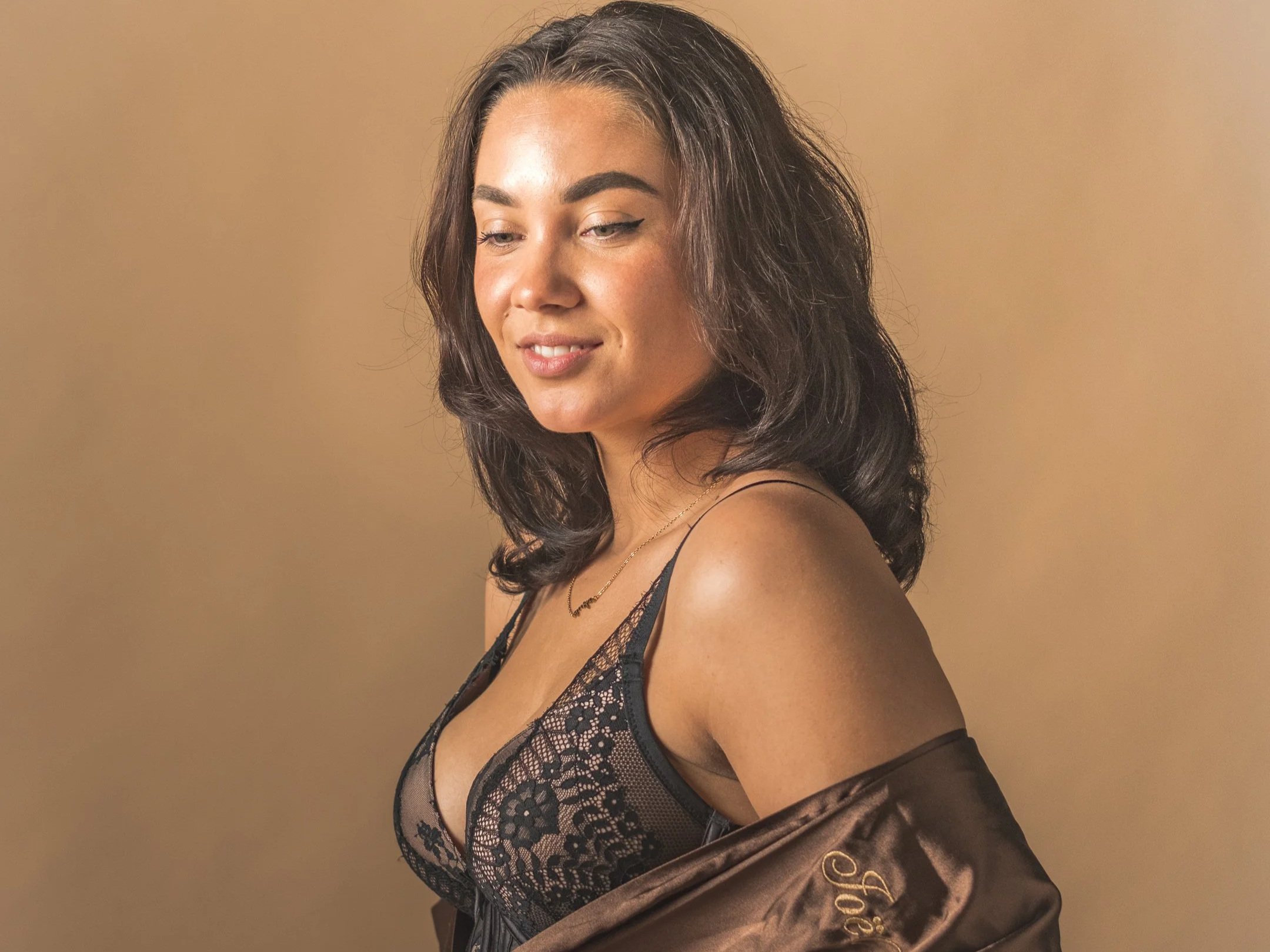 Close-up van een vrouw met bruin haar in een zwarte kanten lingerie, zittend tegen een beige achtergrond, met een satijnen bruin jasje over haar schouder.