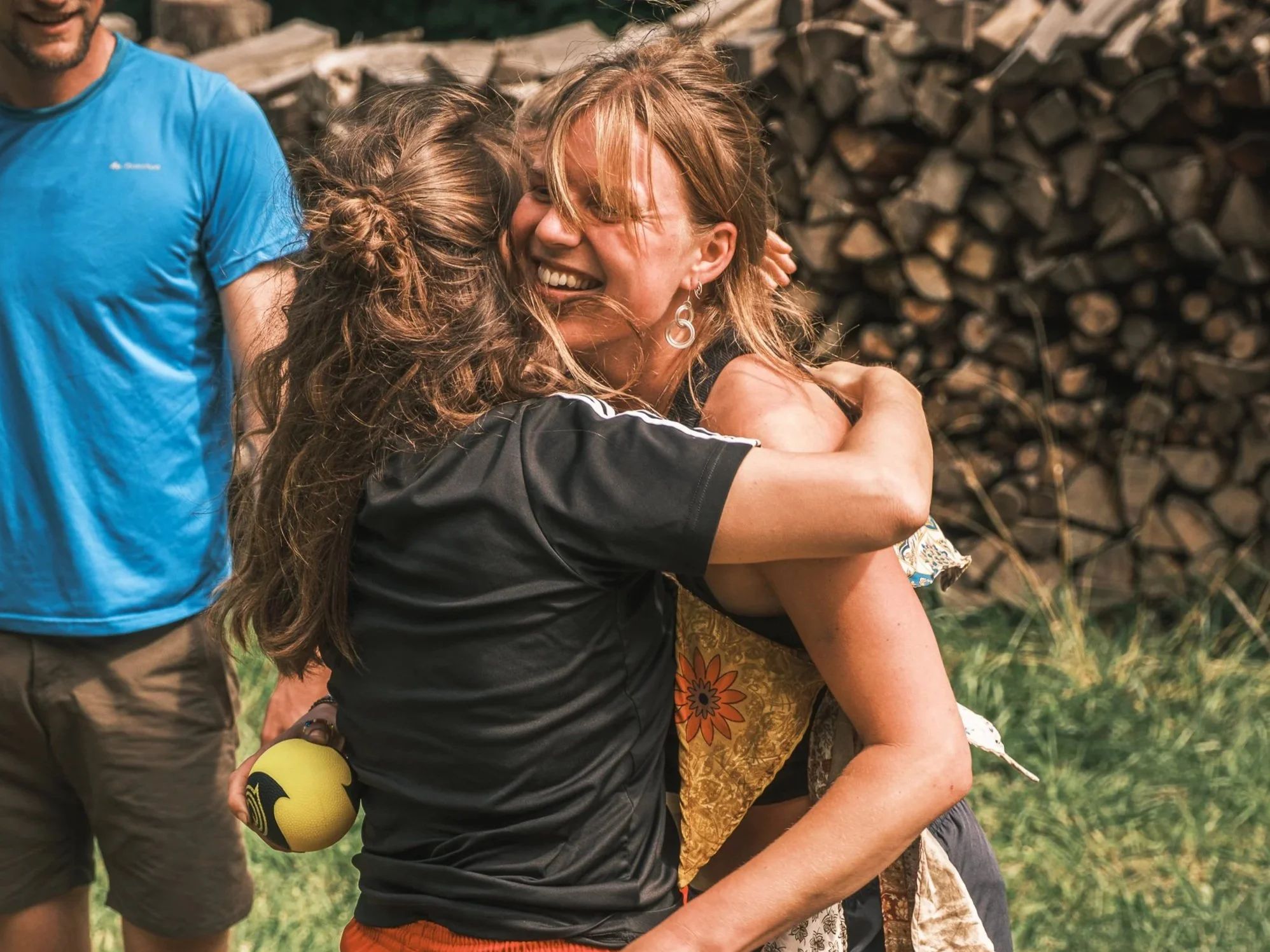 Twee vrouwen omhelsingen, lachend, buiten met houtstapels op de achtergrond.