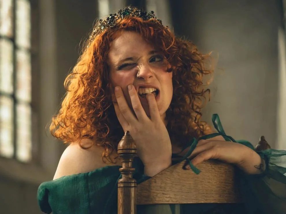 Vrouw met rood krullend haar, glimlachend en knipogend, zittend op een houten stoel met een kroon op haar hoofd en een groene jurk.