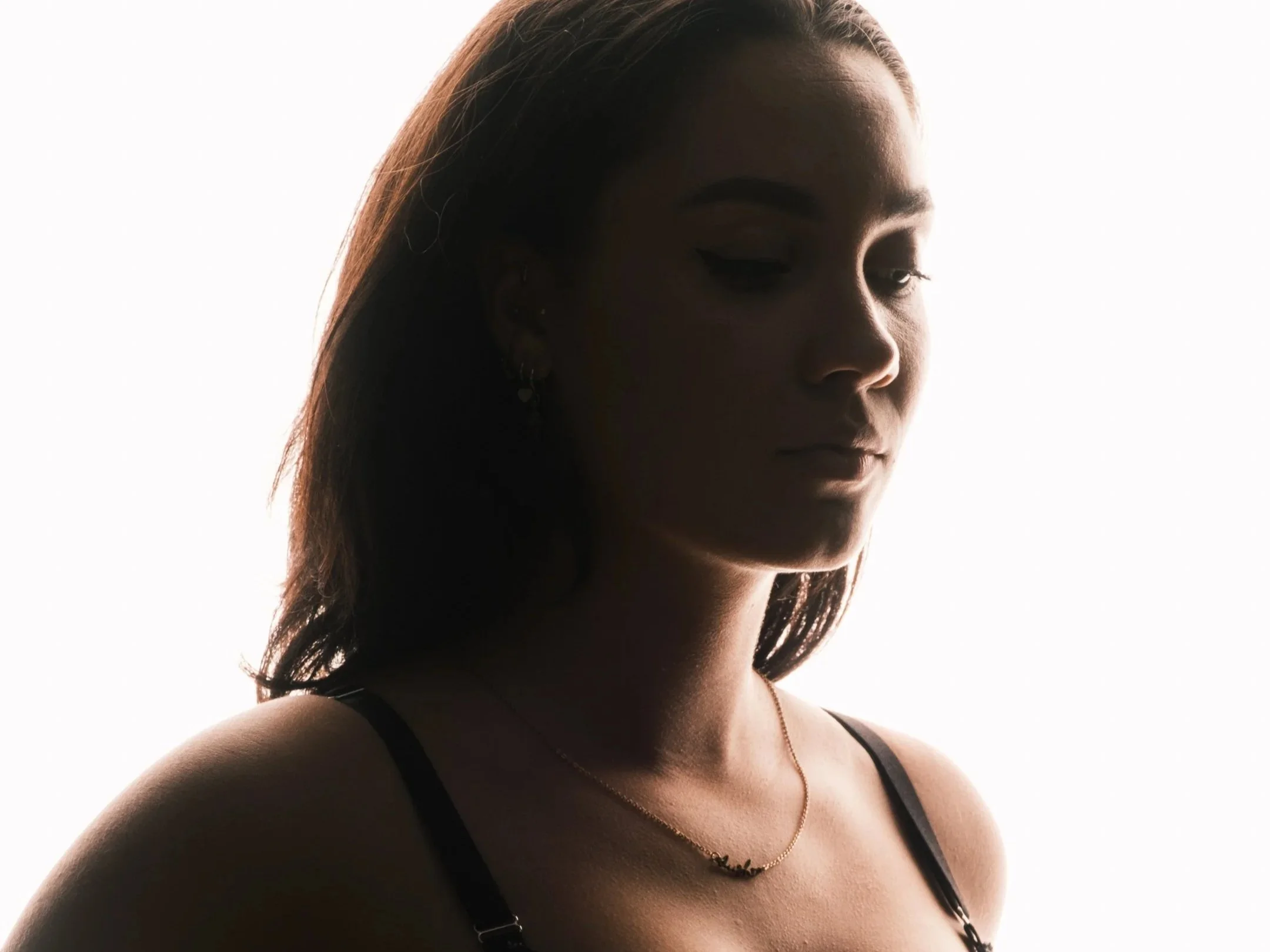 Portret van een jonge vrouw met lang, donker haar, die naar beneden kijkt, gekleed in een zwart topje en een fijne ketting, op de achtergrond dichthelder.