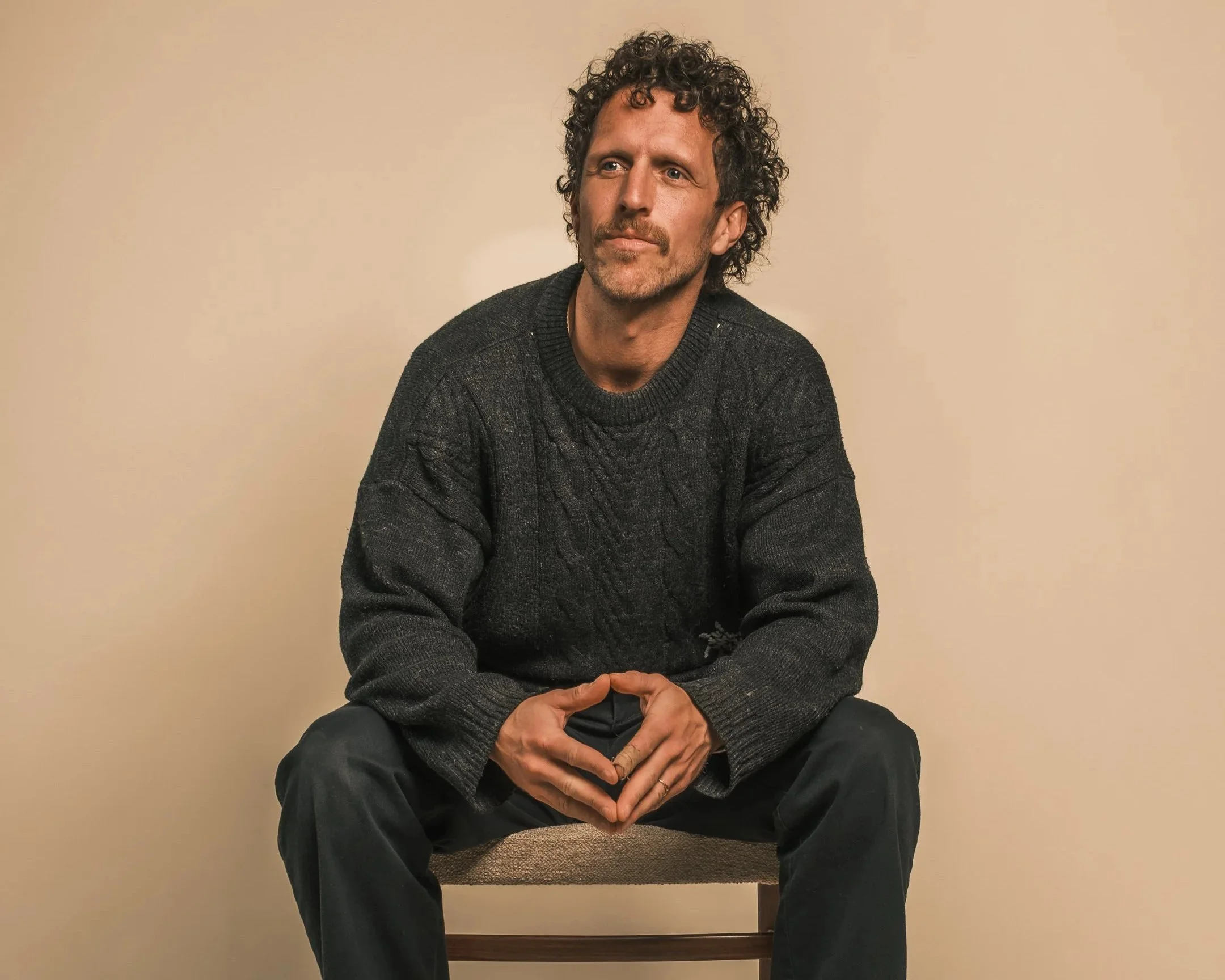 Man met curly haar en een baard, zittend op een stoel, gekleed in een donkergrijze sweater en donkere broek, voor een beige achtergrond.