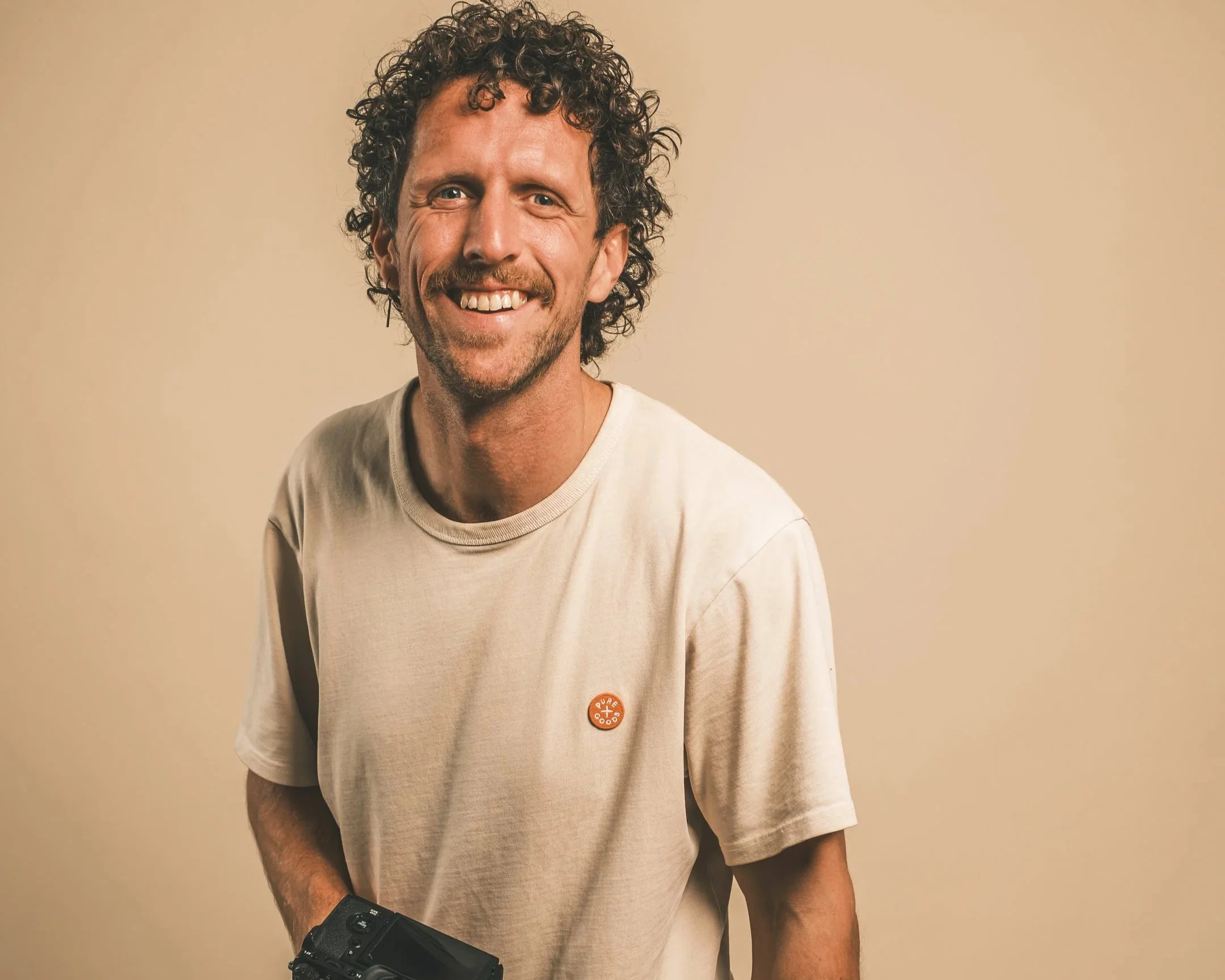 Man met curly haar die lacht en een fotocamera vasthoudt, tegen een effen beige achtergrond.