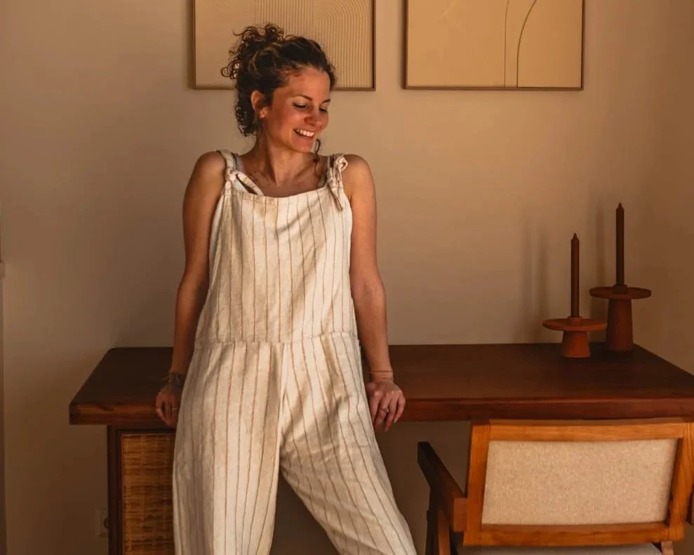 Vrouw met bruin gekruld haar in een spijkerbroek en top, glimlachend in een interieur met houten meubelen en moderne kunst aan de muur.