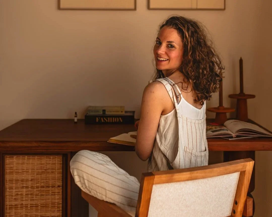 Vrouw zittend op een stoel bij een bureau, lachend en naar de camera kijkend, in een kamer met wanden die versierd zijn met twee schilderijen.
