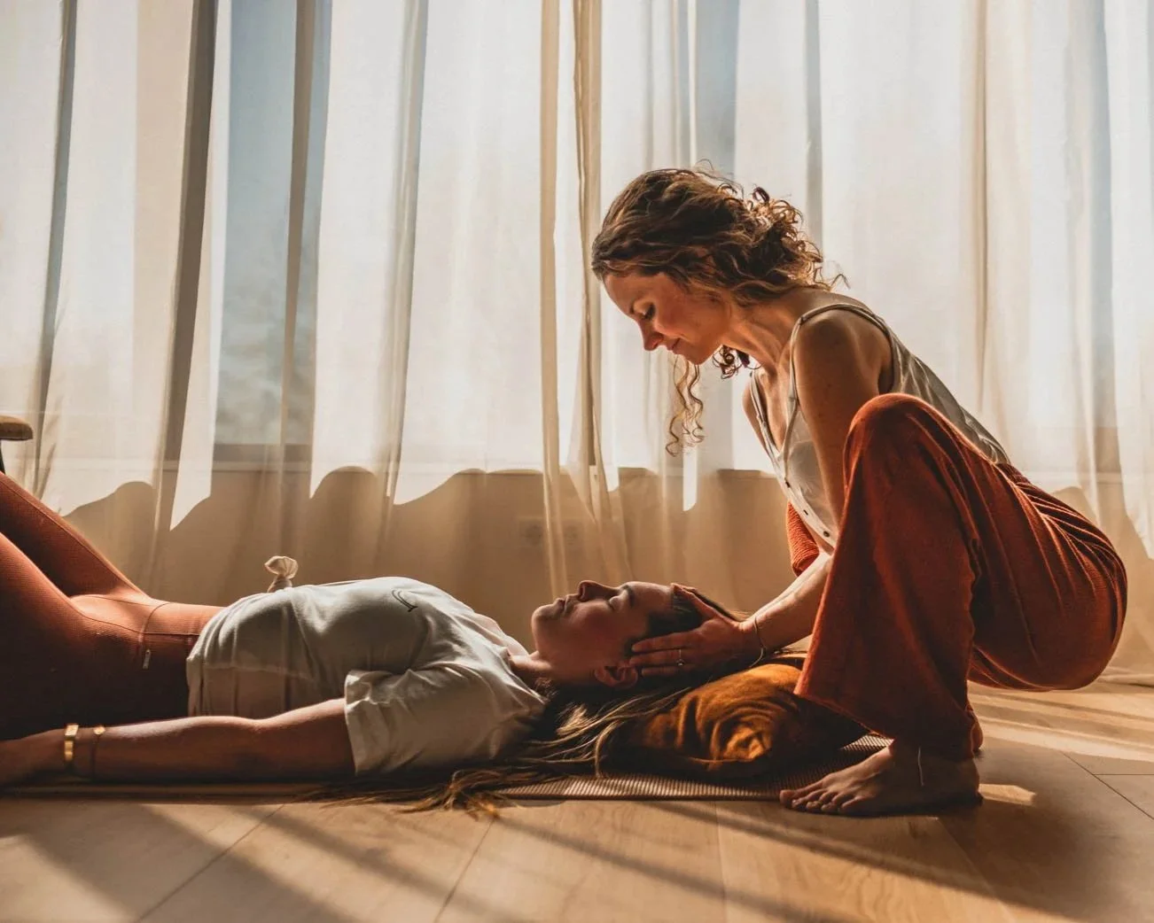 Een vrouw ligt op de vloer met gesloten ogen, terwijl een andere vrouw knielt naast haar en haar hoofd ondersteunt.