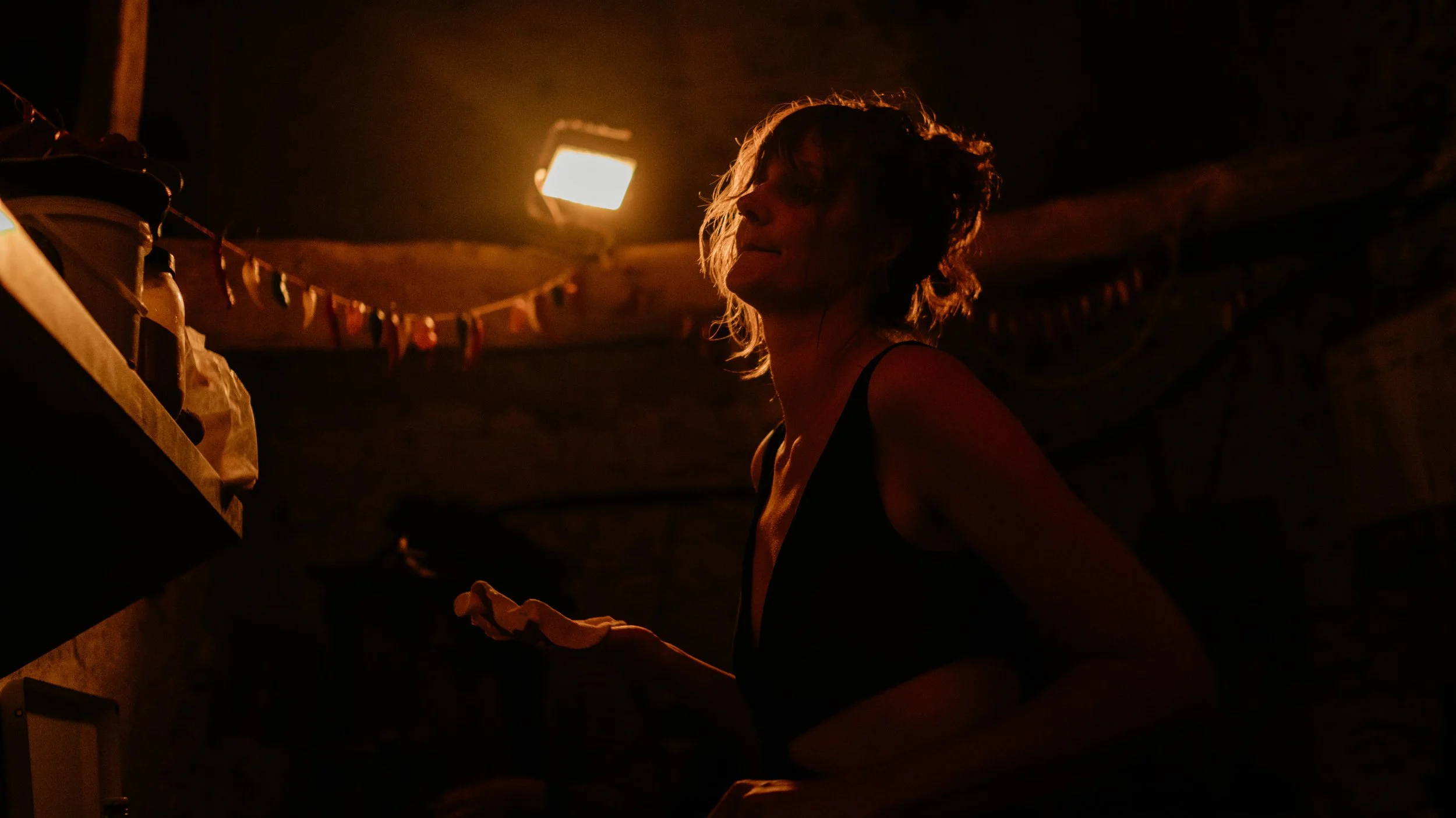 Een vrouw met wit haar die in een donkere kamer staat onder een warm licht, met haar ogen dicht en een hand omhoog terwijl ze iets vasthoudt.