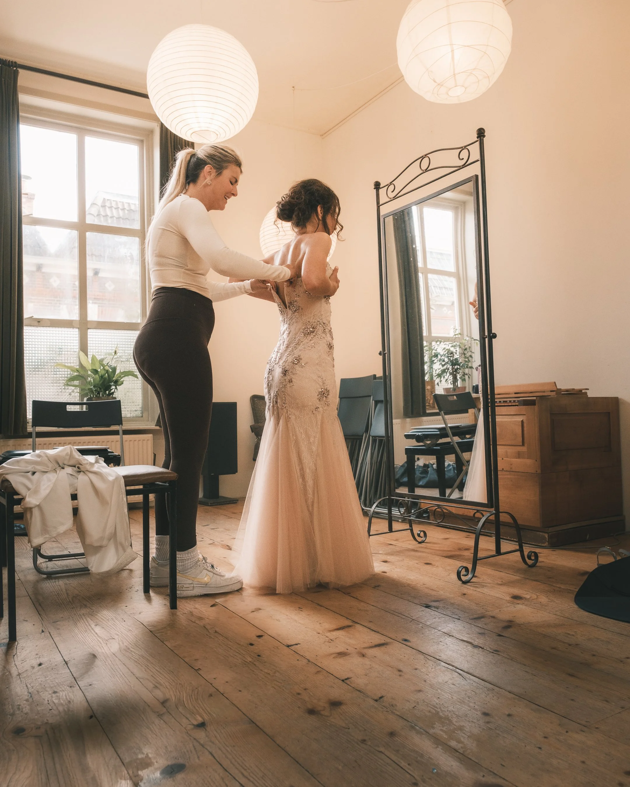 Vrouw helpt bruid zich te kleden in een kamer met houten vloer, grote ramen en een grote spiegel.