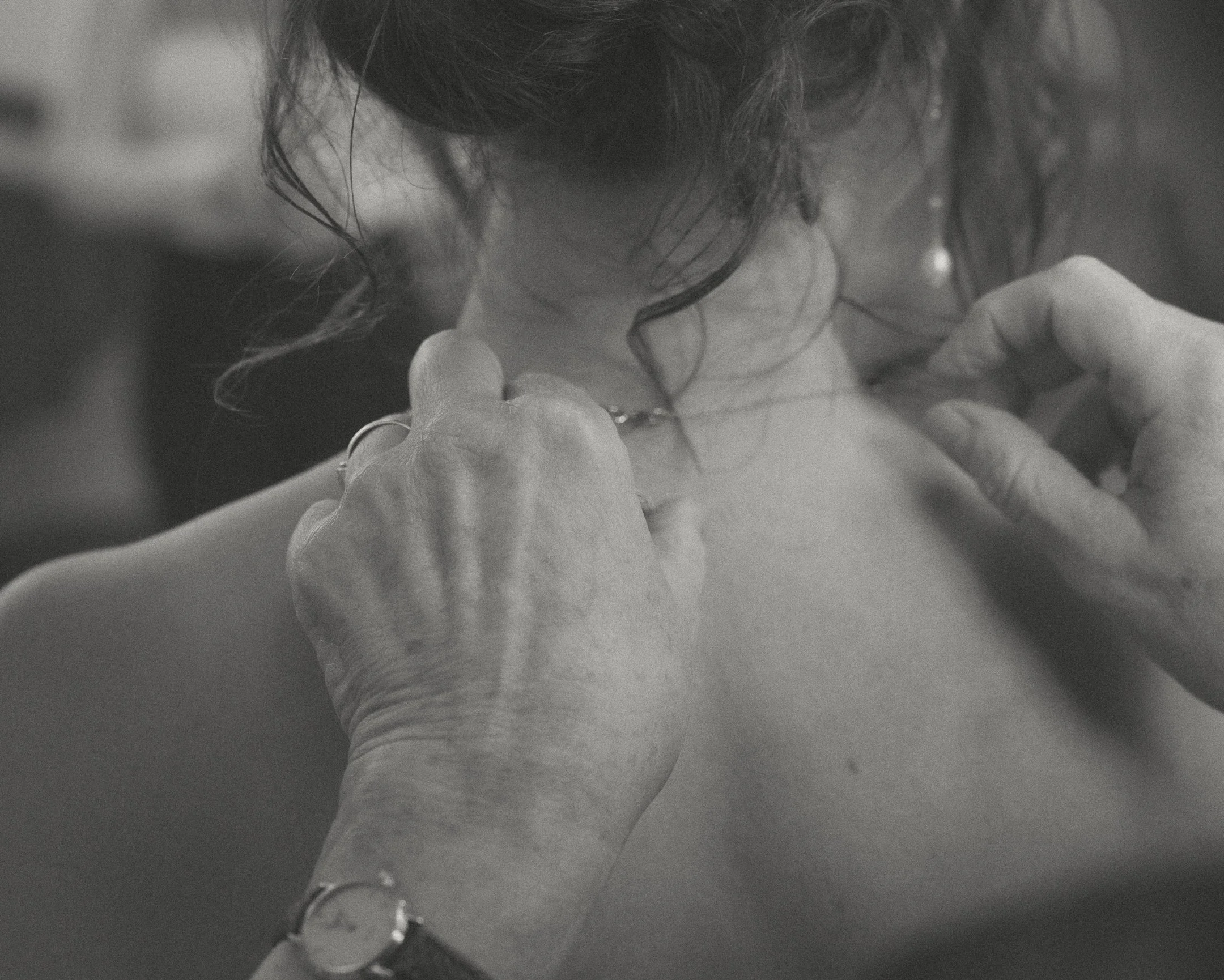 Close-up zwart-wit foto van een persoon die lichtjes wordt gekleed door een ander persoon, met de nadruk op handen en de nek, in een intieme en delicate setting.