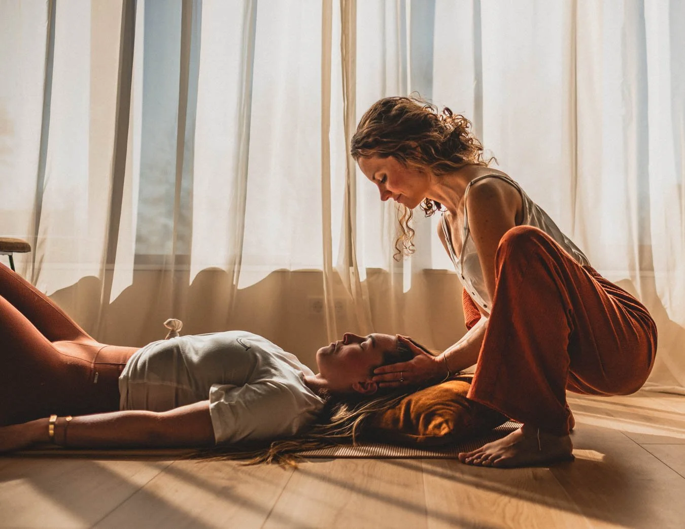 Twee vrouwen in een kamer met zacht licht, een liggend op de grond met hoofd op een kussen en een zittende vrouw die haar helpt. Het lijkt een moment van emotionele ondersteuning of meditatie.