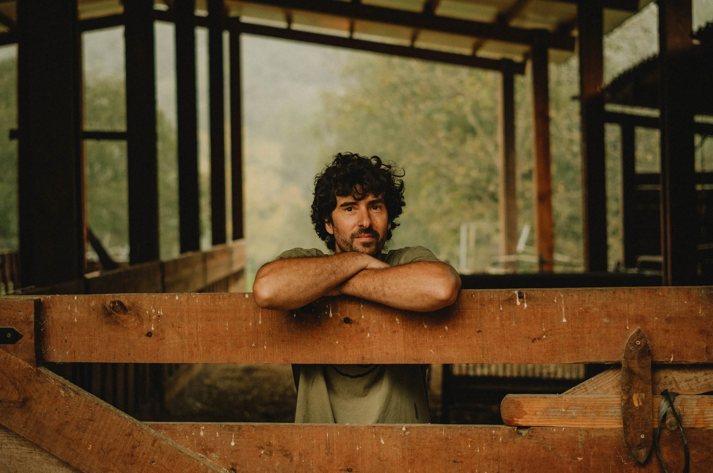 Man met donkere, curly haar rust op een houten schutting in een houten stal of schuur, met een groene achtergrond van bomen.