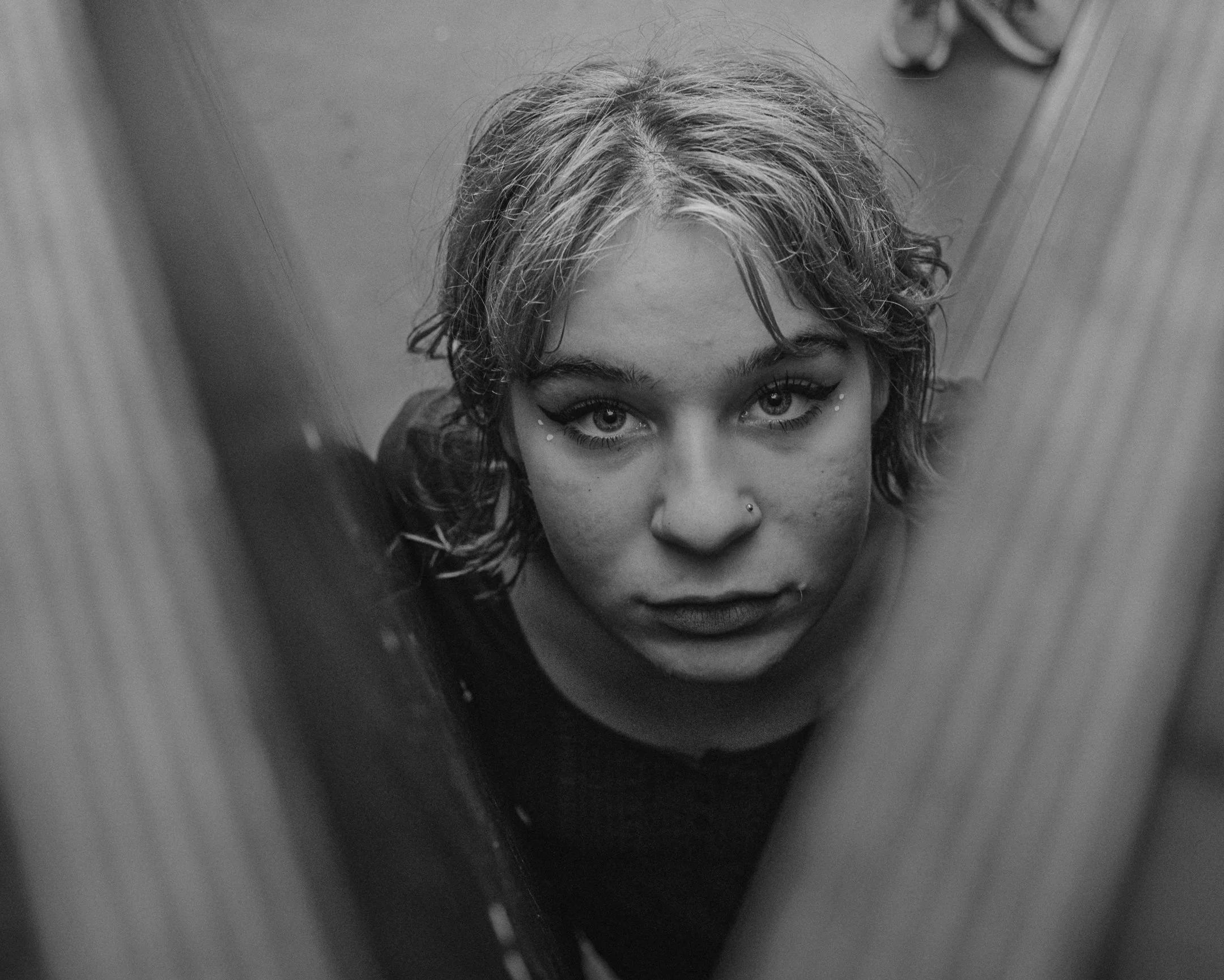 Een jonge vrouw kijkt omhoog in de camera, haar gezicht tussen houten panelen, in zwart-wit.