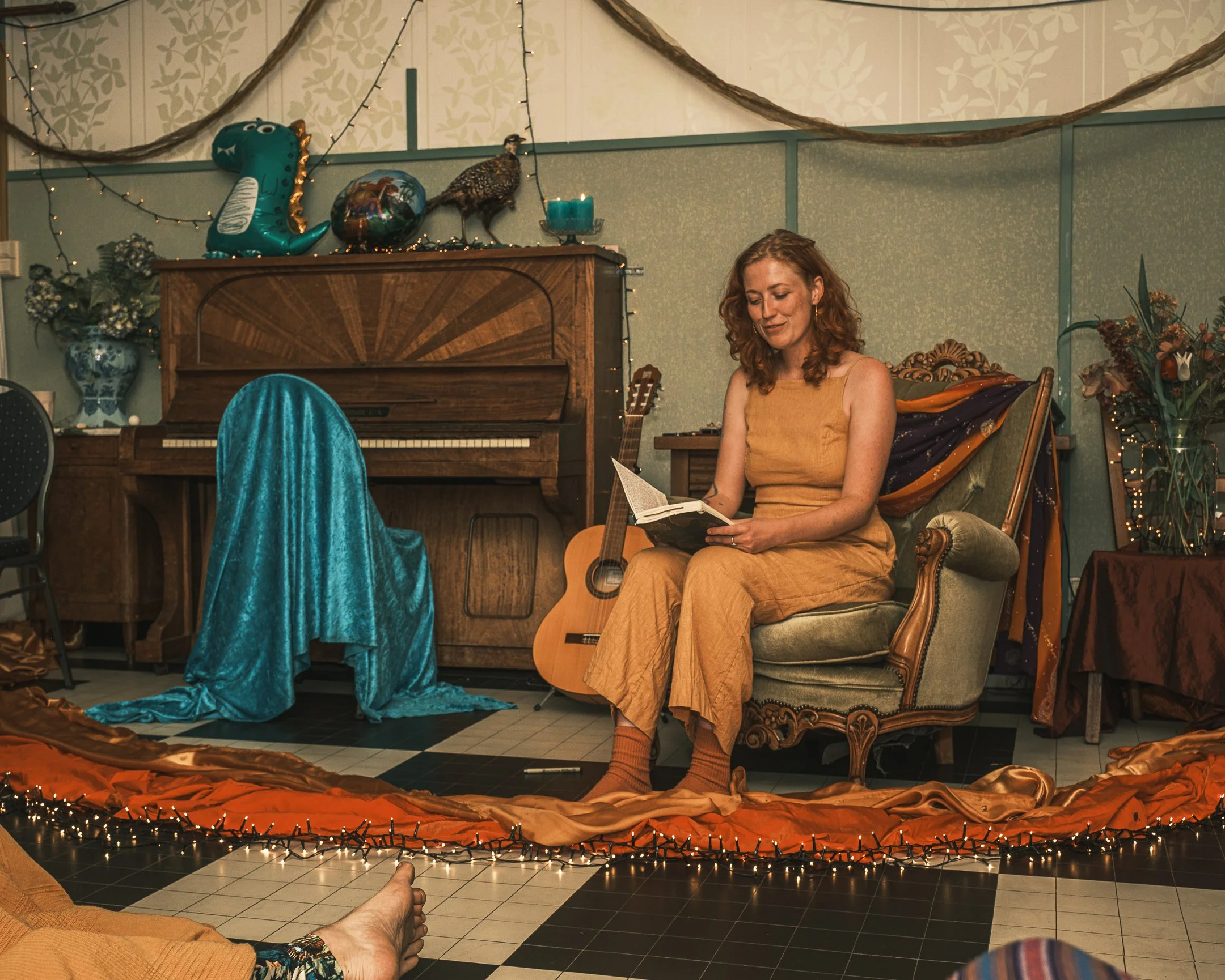 Een vrouw zit in een vintage fauteuil, leest een boek, op een feestelijke setting met decoraties, onder andere een piano, gitaar en ballonnen.