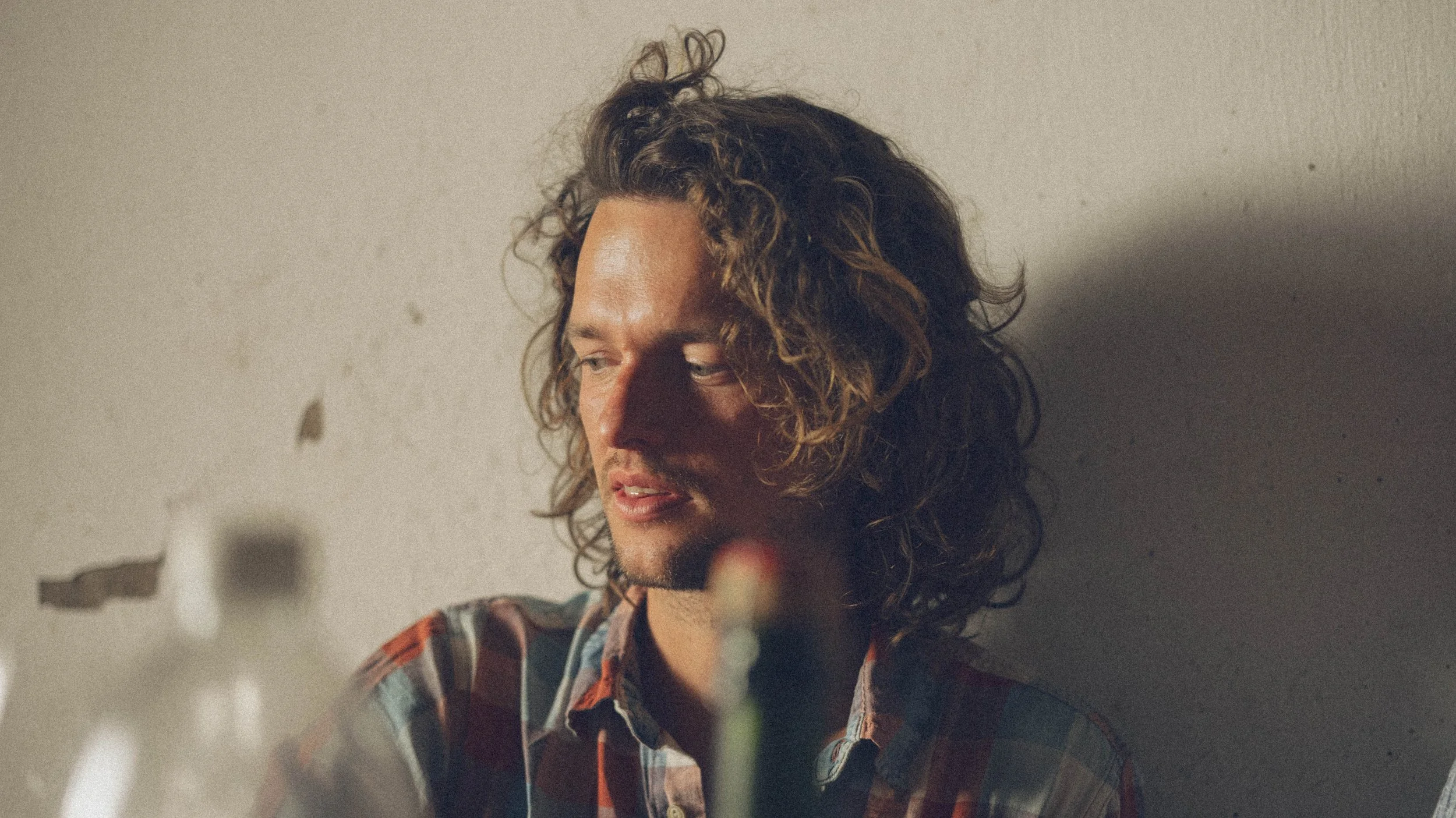 Een jonge man met golvend bruin haar die naar links kijkt, zittend tegen een muur met een kleine beschadiging, met een schemerige sfeer en zachte verlichting.