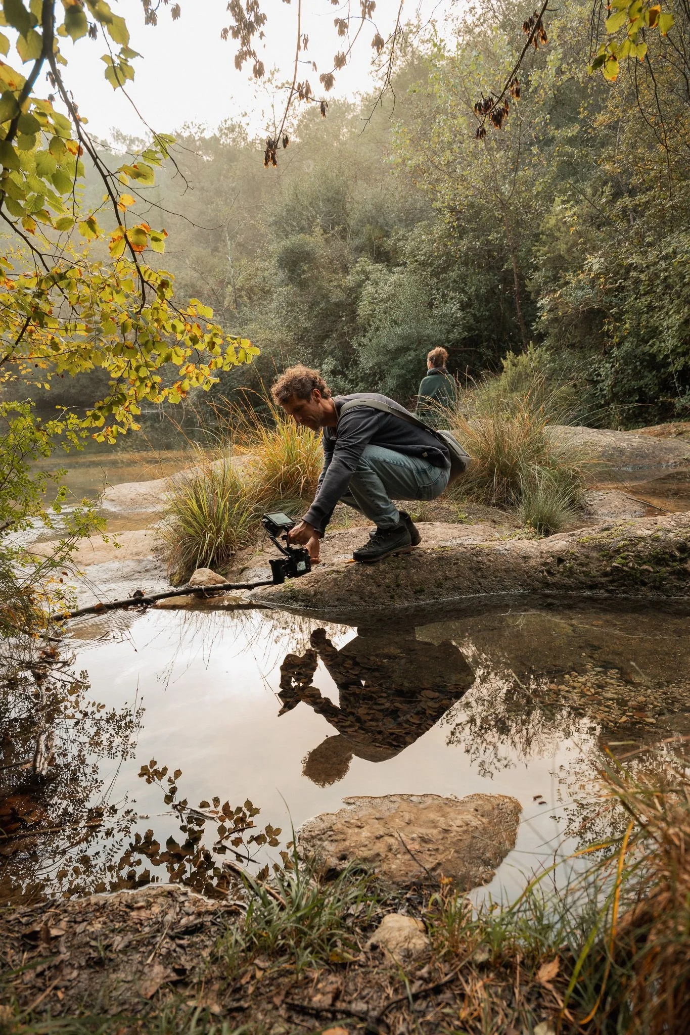 Man die een camera op een takje boven een waterpartij plaatst terwijl er een vrouw in de achtergrond kijkt.