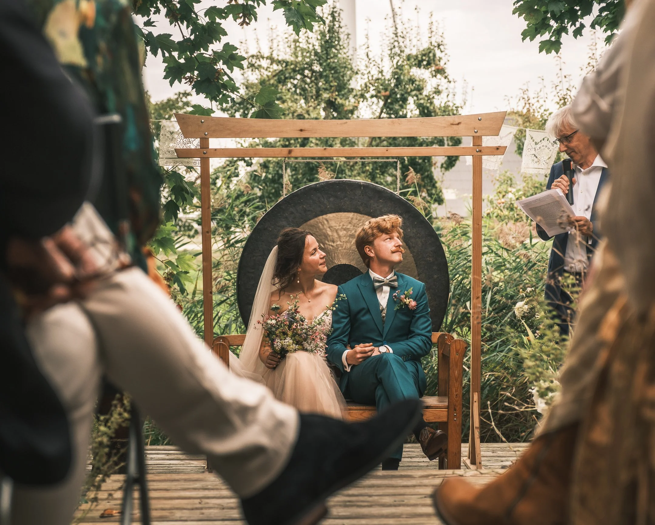 Een bruid en bruidegom zitten op een bankje tijdens een huwelijk in de tuin, omringd door vrienden en familie, met een spreker in de rechterbovenhoek die een speech houdt.