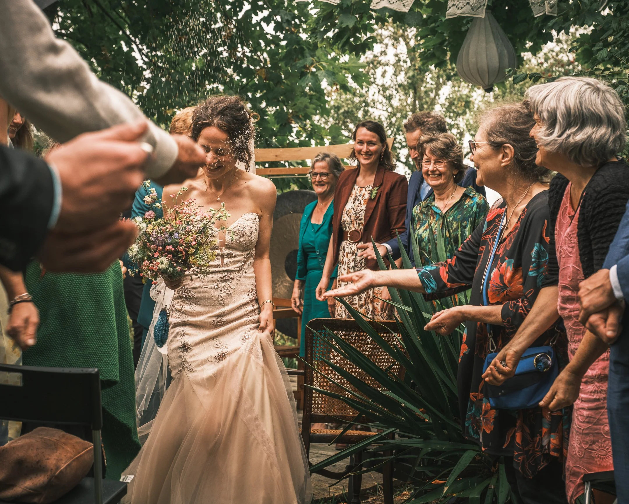 Wedding ceremony outdoors with a bride, dressed in a strapless wedding gown holding a bouquet, and gathered guests smiling and celebrating under trees.