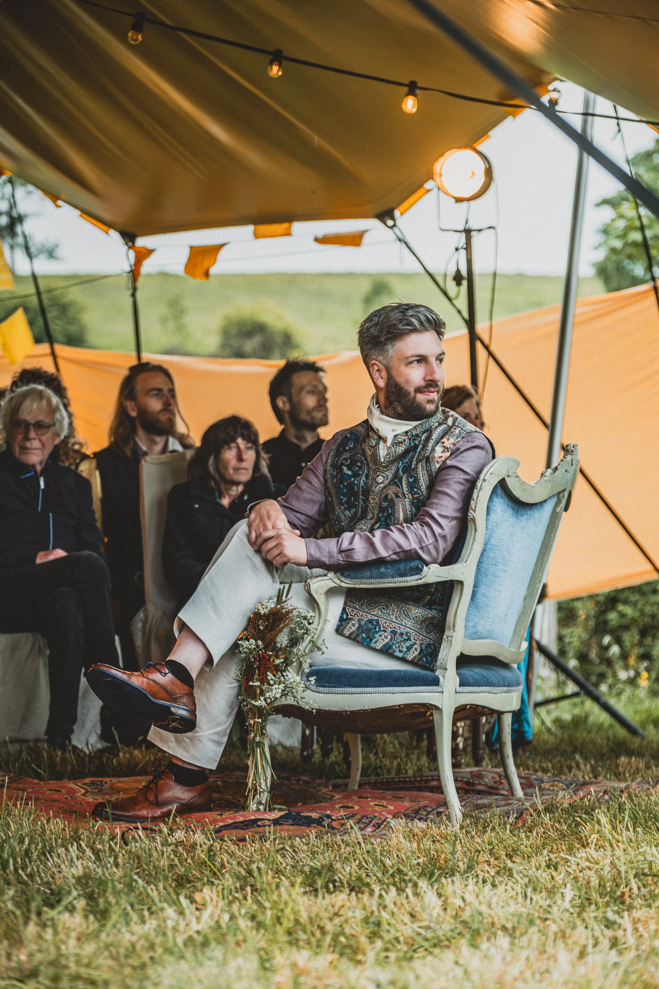 Man zit op een vintage blauwe fauteuil onder een tent met gele en oranje decoraties, omringd door luisterende mensen, tijdens een buitenevenement.