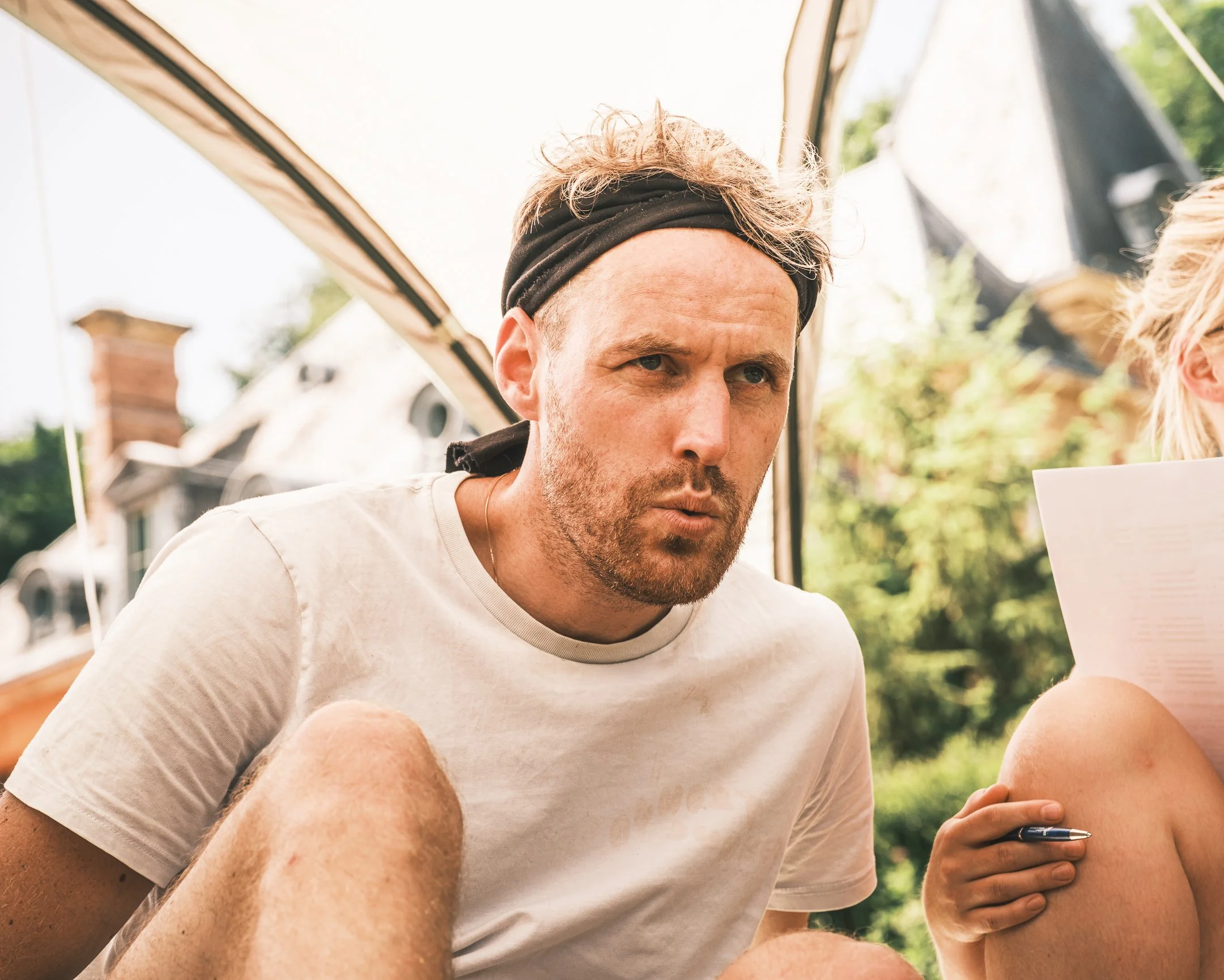 Man wearing a black headband, white t-shirt, looking serious, sitting outdoors under a canopy with blurred trees and house in background.