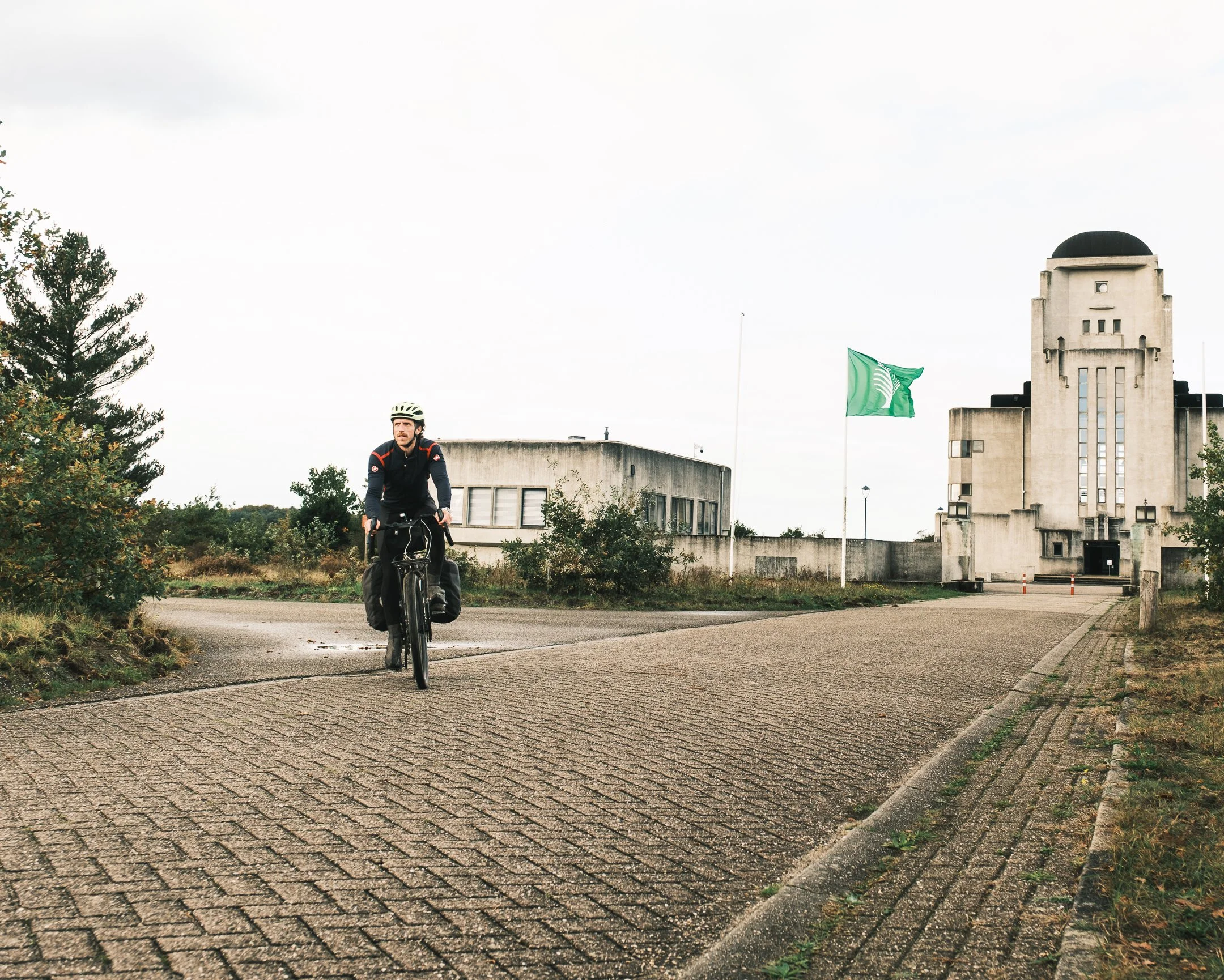 Man fietst op een pad met een historisch gebouw op de achtergrond, omgeven door struiken en een vlag met groene en witte strepen.