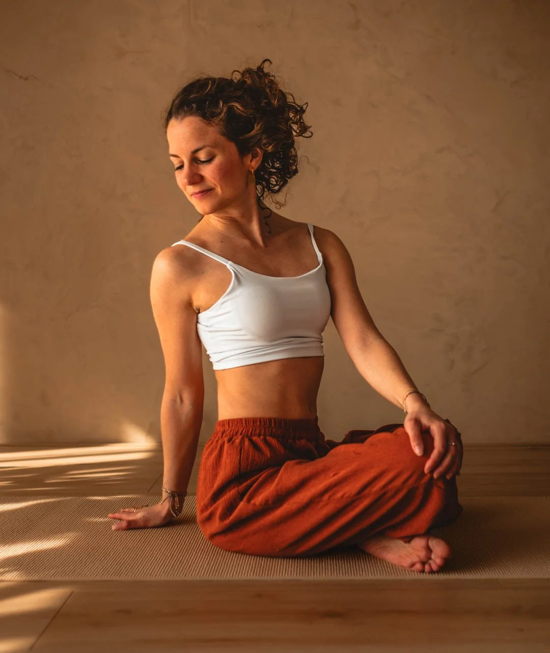 Een vrouw zit in een yogapose op een mat, in een rustige omgeving met beige muren.