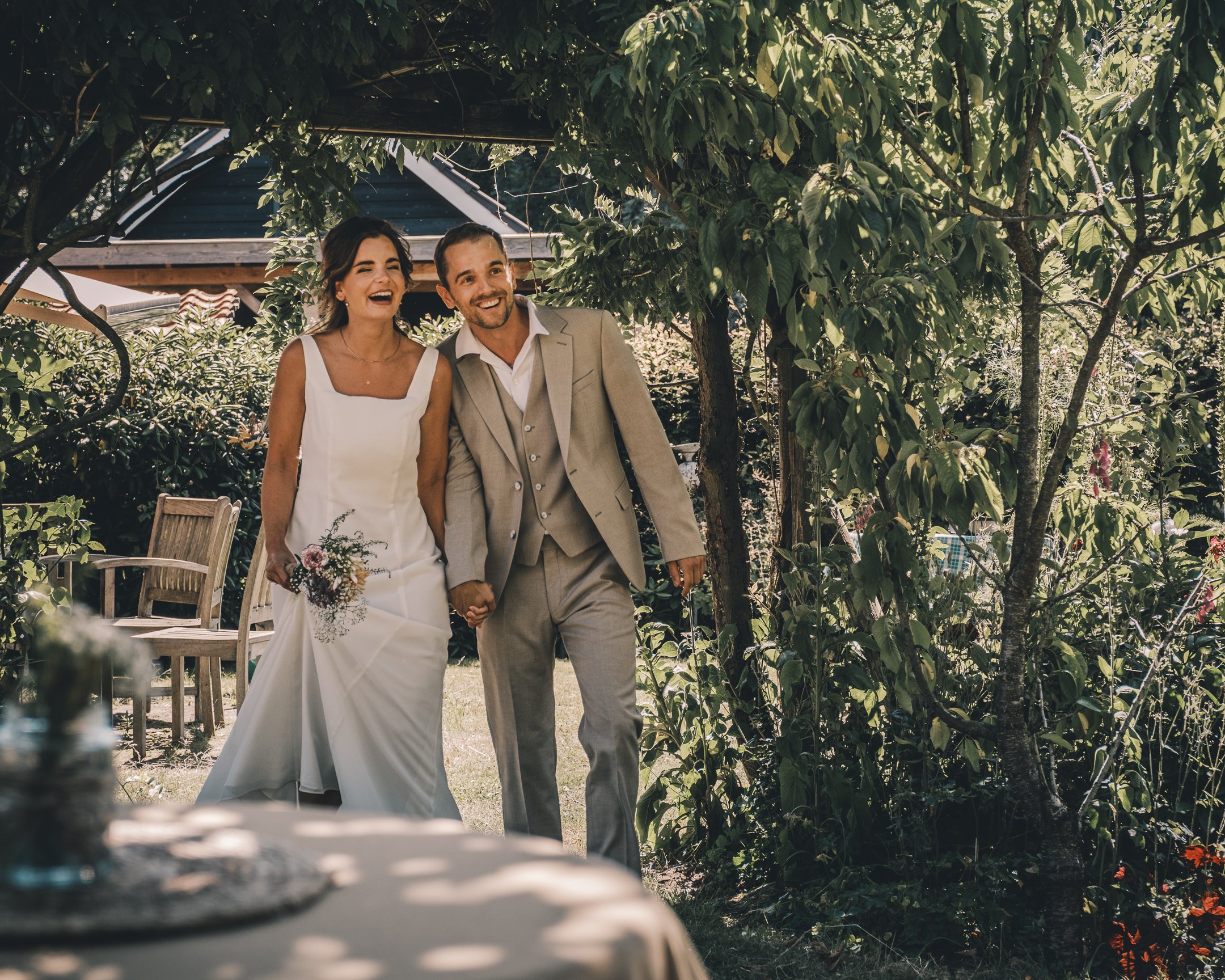 Bruid en bruidegom lopen hand in hand door een tuin tijdens hun huwelijksfeest, lachend en genietend van de dag.