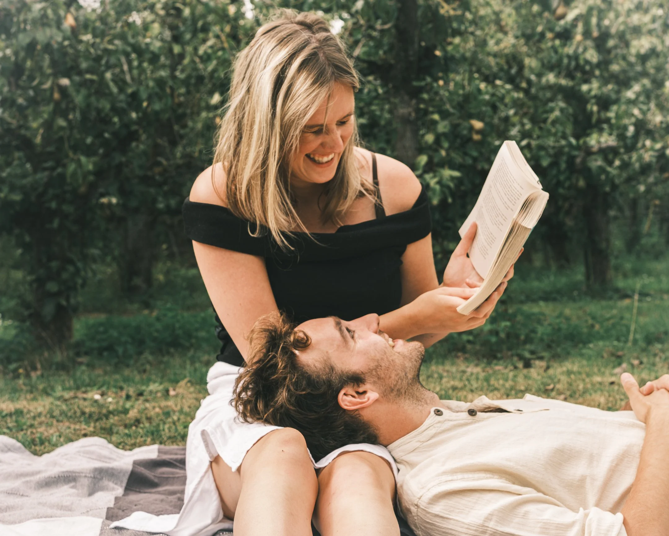 Een lachende vrouw leest een boek voor een man die op de grond ligt in een park.