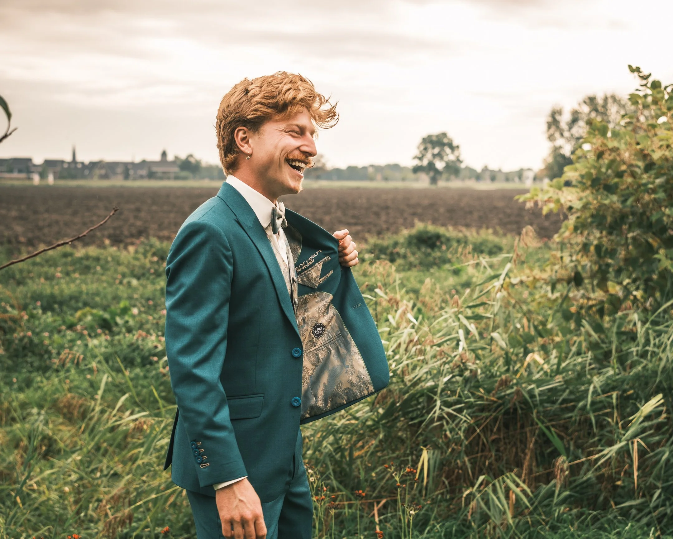 Man in een groene maatpak staat in een weiland met groen gras en struiken, lachend terwijl hij zijn jas openspert. Op de achtergrond zijn akkers en bomen zichtbaar onder een bewolkte hemel.