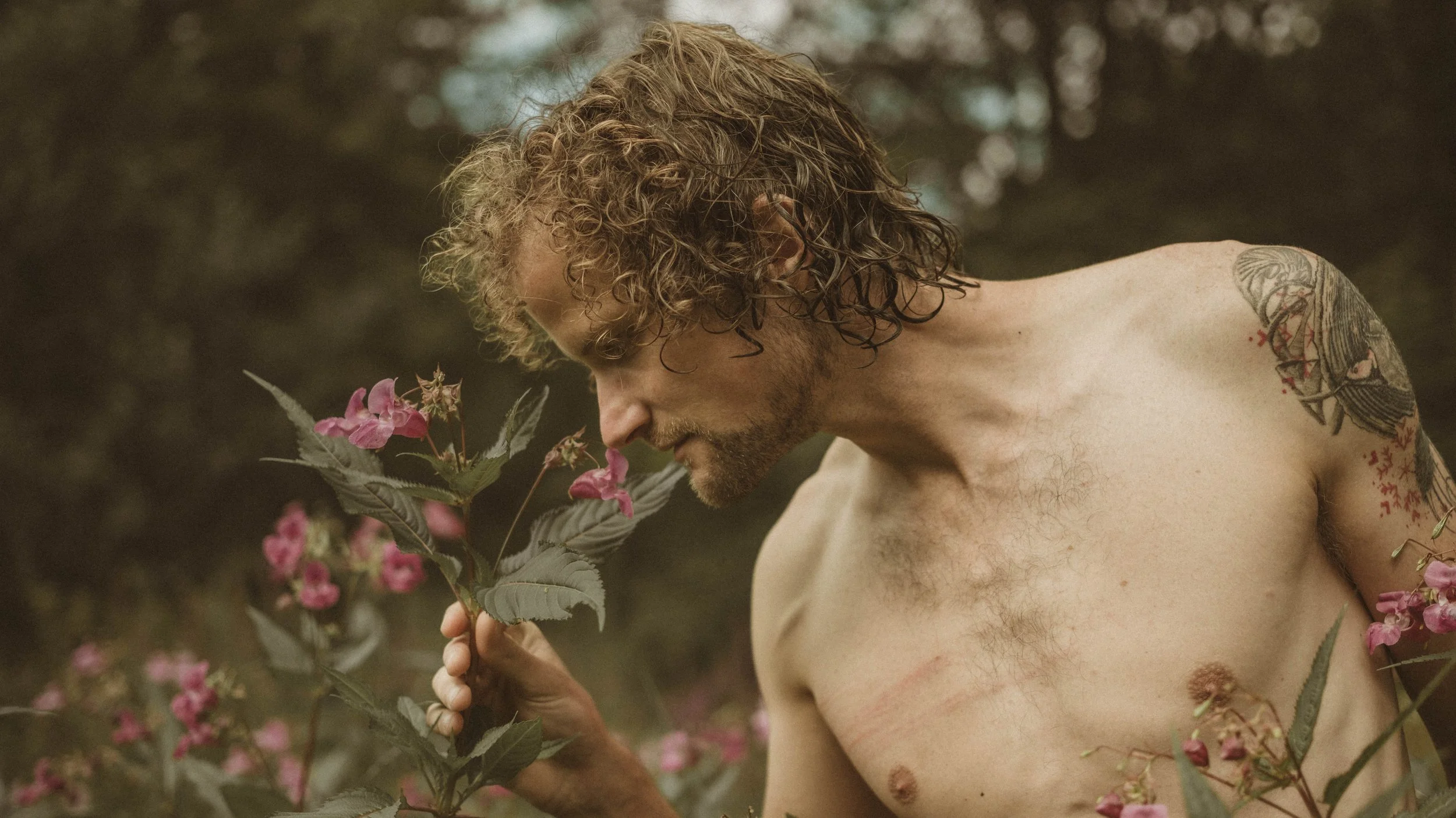 Een man met lang, gekruld haar en tattoos op zijn schouder en arm, die ruikt aan roze bloemen in een natuurlijk landschap.