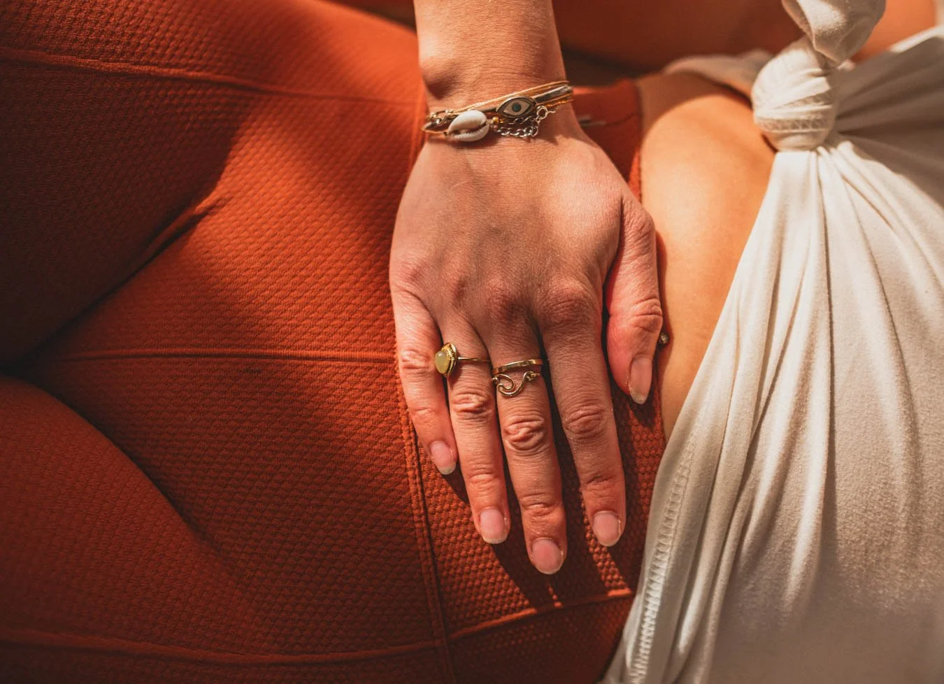Close-up van een hand met sieraden rustend op een oranje stoffen oppervlak, met een witte stof met knoopzak zichtbaar.