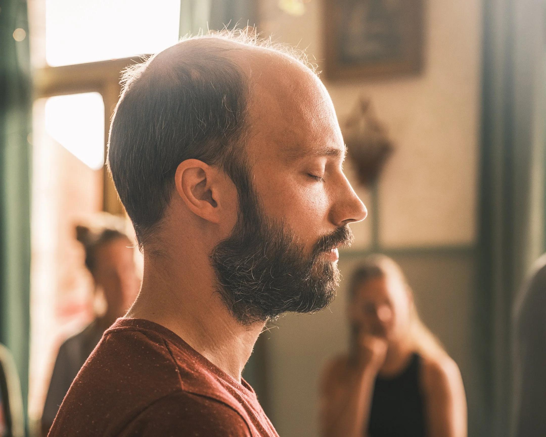 Man met gesloten ogen in meditatie, binnen met natuurlijke lichtinval en mensen op de achtergrond.