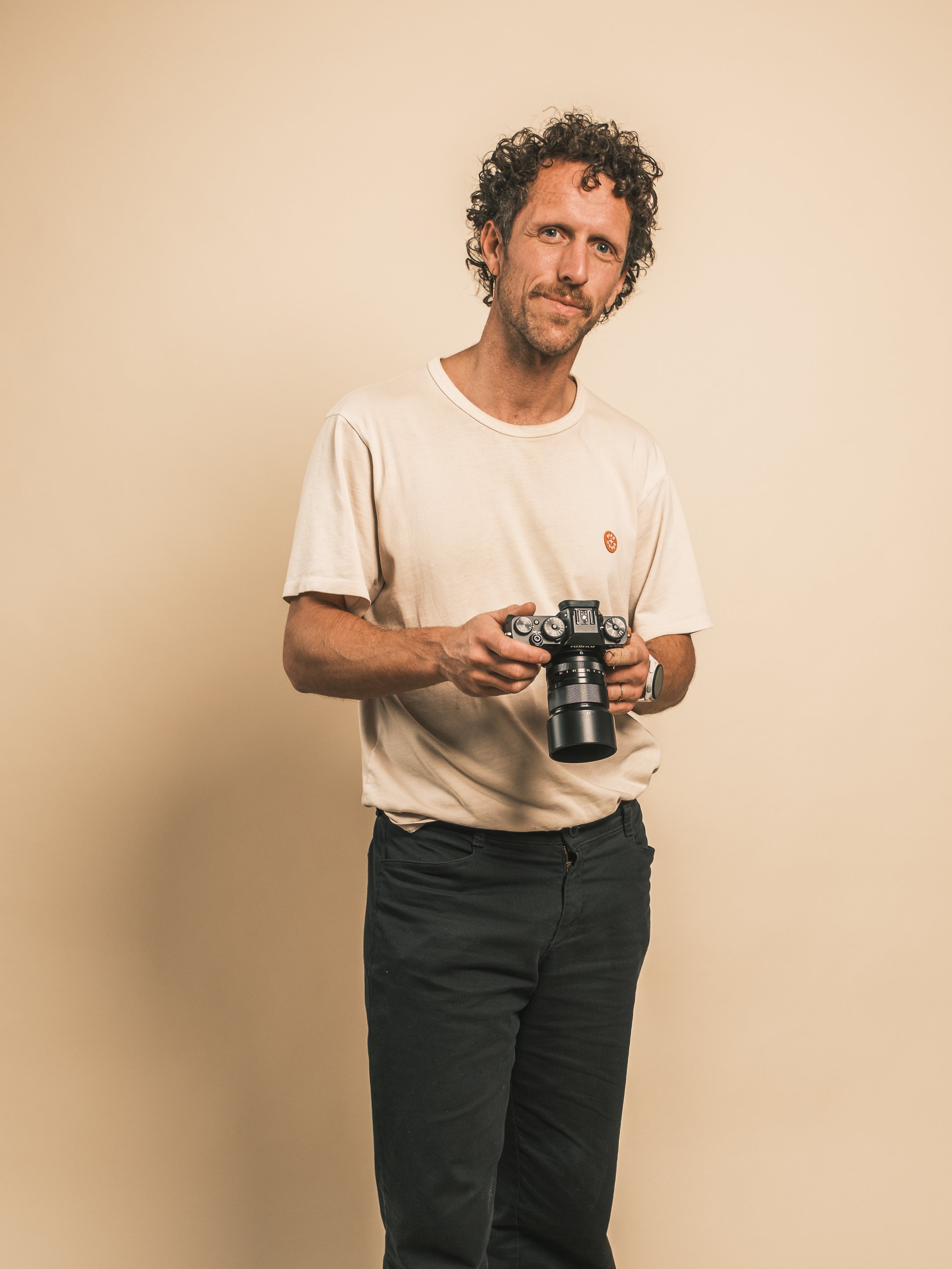 Man met verwassen witte T-shirt en zwarte broek die een camera vasthoudt, staand voor een effen beige achtergrond.
