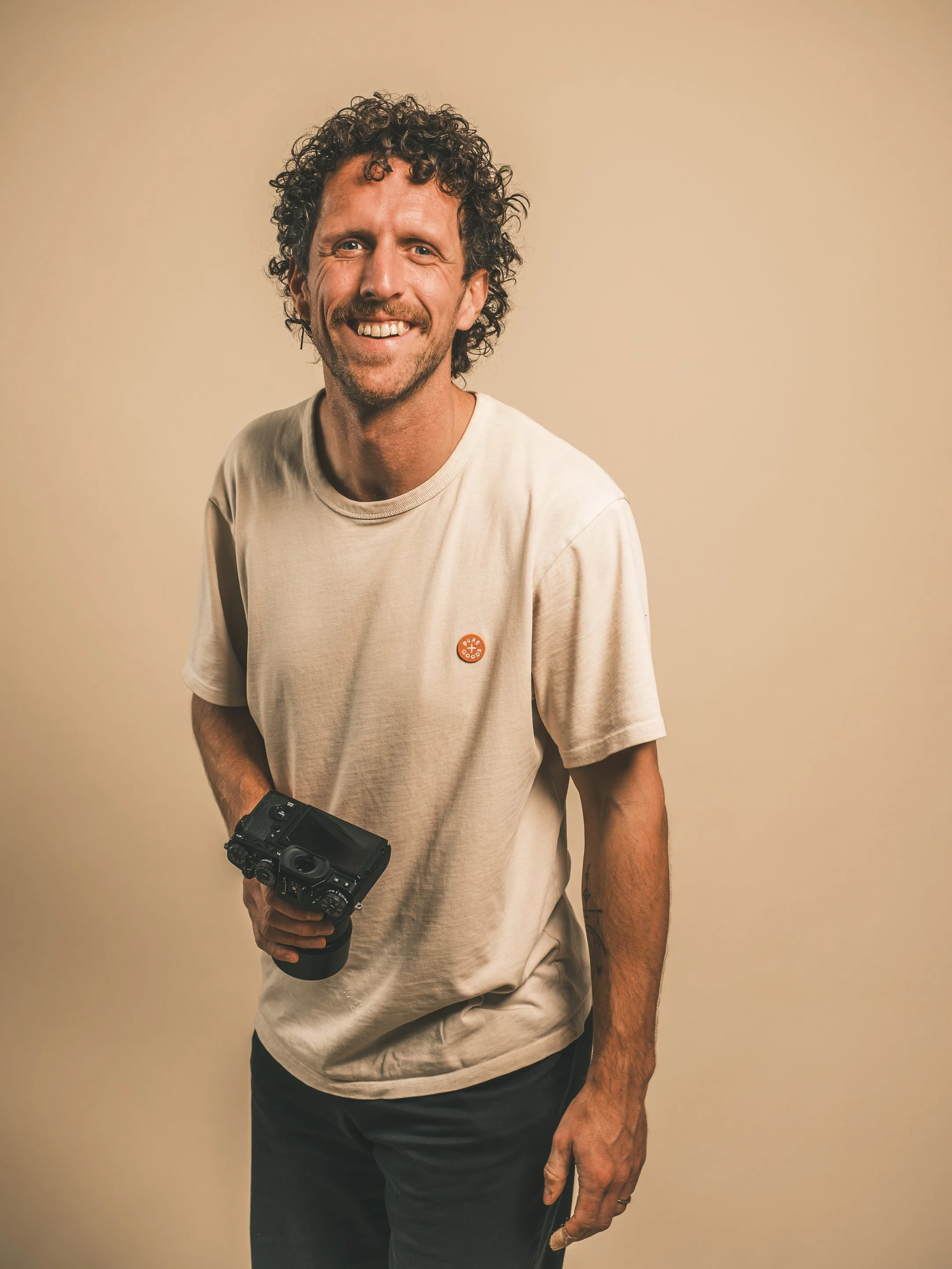 Man met curly haar en glimlach, wit T-shirt met logo, holdende camera, staand voor een effen achtergrond.