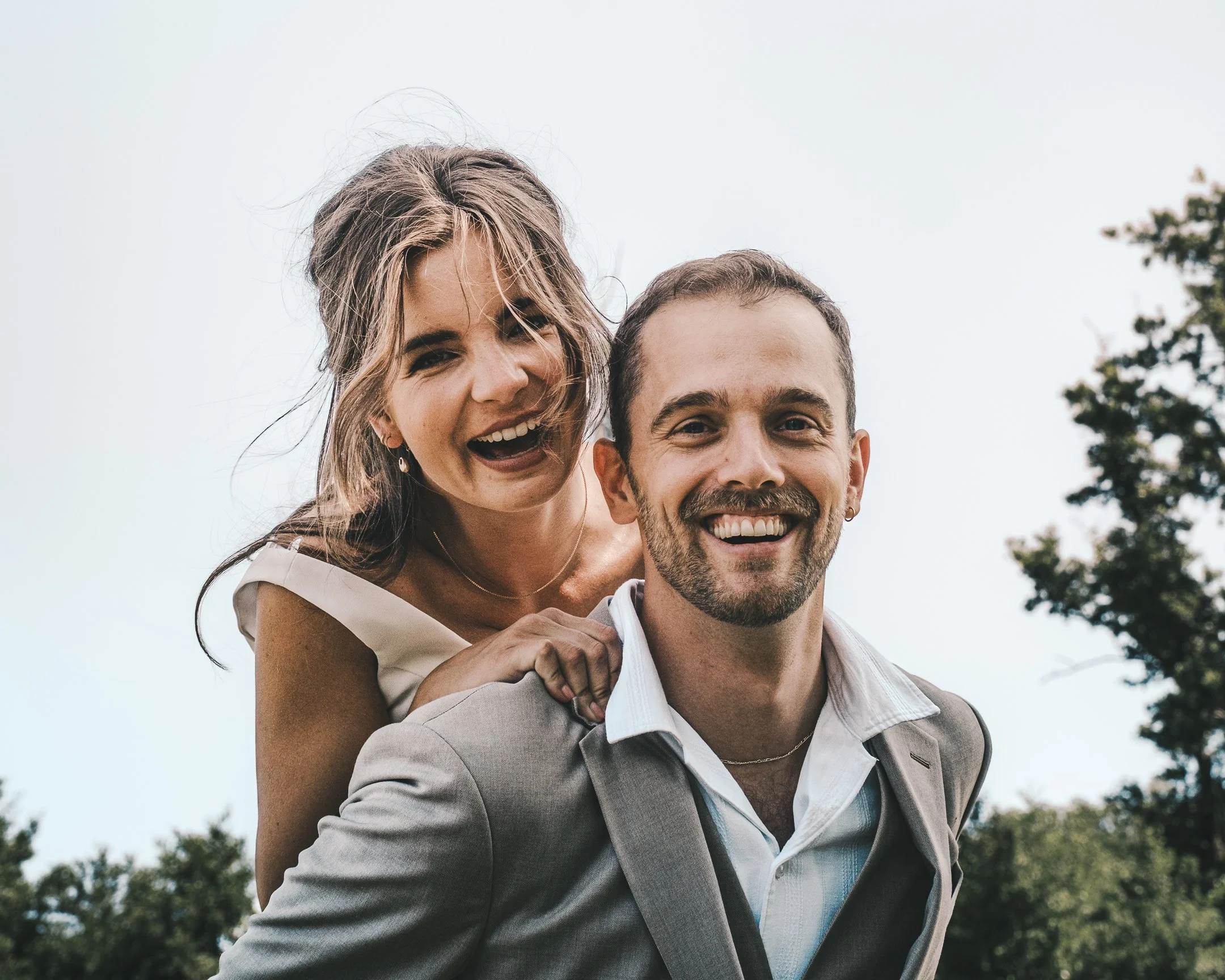 Een blije man in een grijs pak met wit overhemd draagt een lachende vrouw met lang haar op zijn rug. Luchten en bomen op de achtergrond.