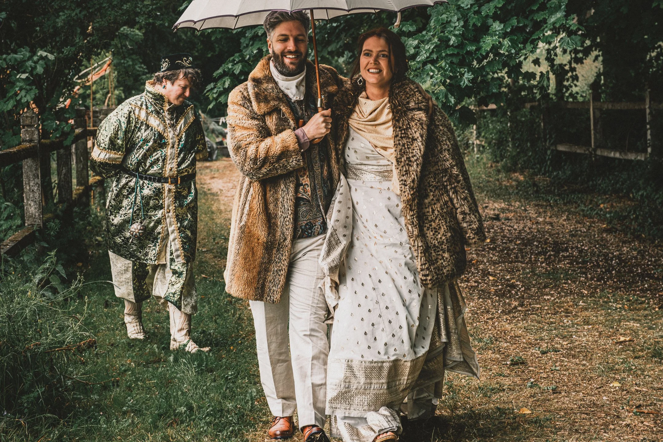Twee mensen die samen lopen op een modderige pad in een bos, lachend onder een paraplu, gekleed in feestelijke en luxe kleding, terwijl een derde persoon op de achtergrond kijkt.