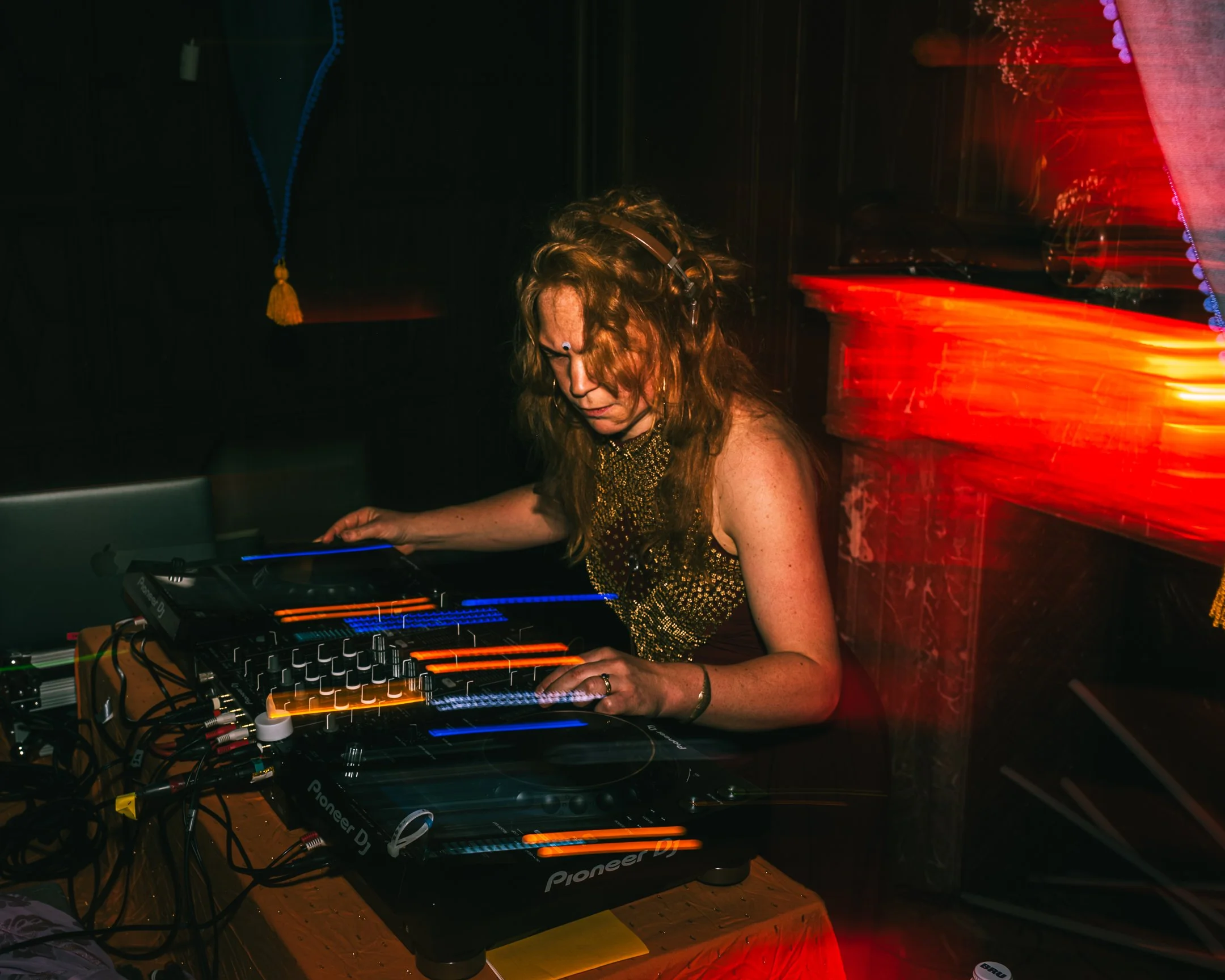 Vrouw met lang, krullend rood haar en koptelefoon, die draait met een DJ-controller in een donkere club, met rode en blauwe lichteffecten.