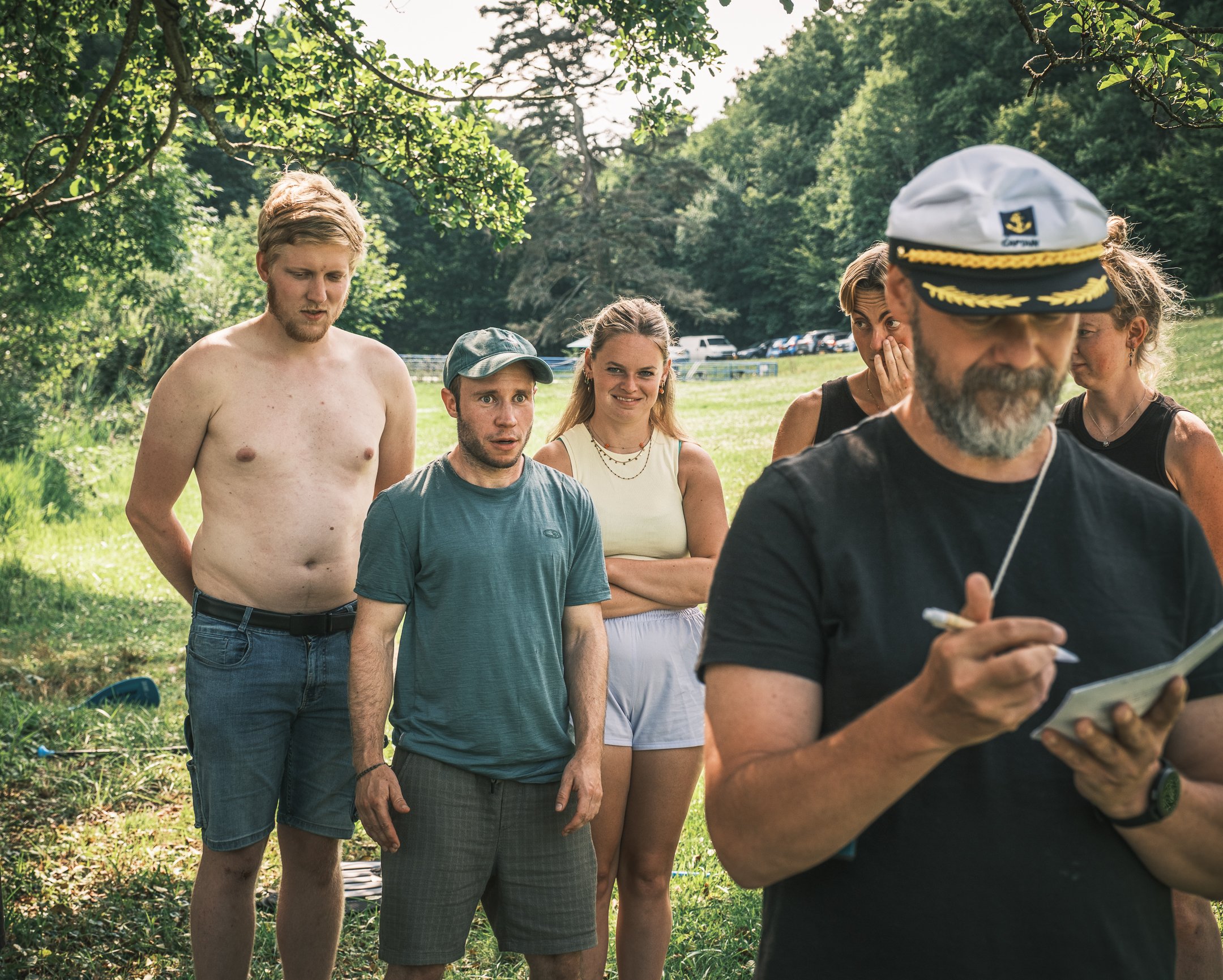 Grupp mensen in park, man met pet schrijft op notitieblok, omringd door vier personen, groene bomen en gras, autos in achtergrond.