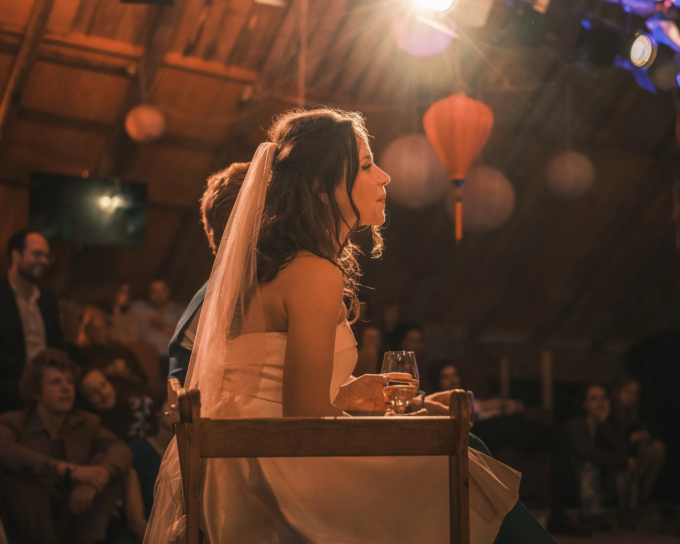 Bride sitting on a wooden chair, holding a glass of wine, with her eyes closed, during a wedding reception in a warmly lit barn decorated with hanging lanterns.