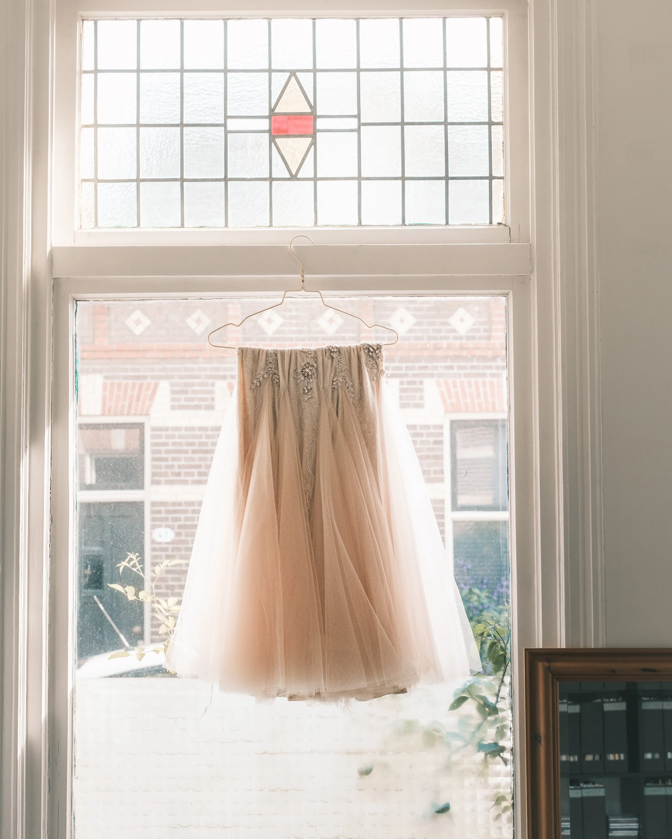 A wedding dress hanging on a wire hanger in front of a window with stained glass and brick building outside.
