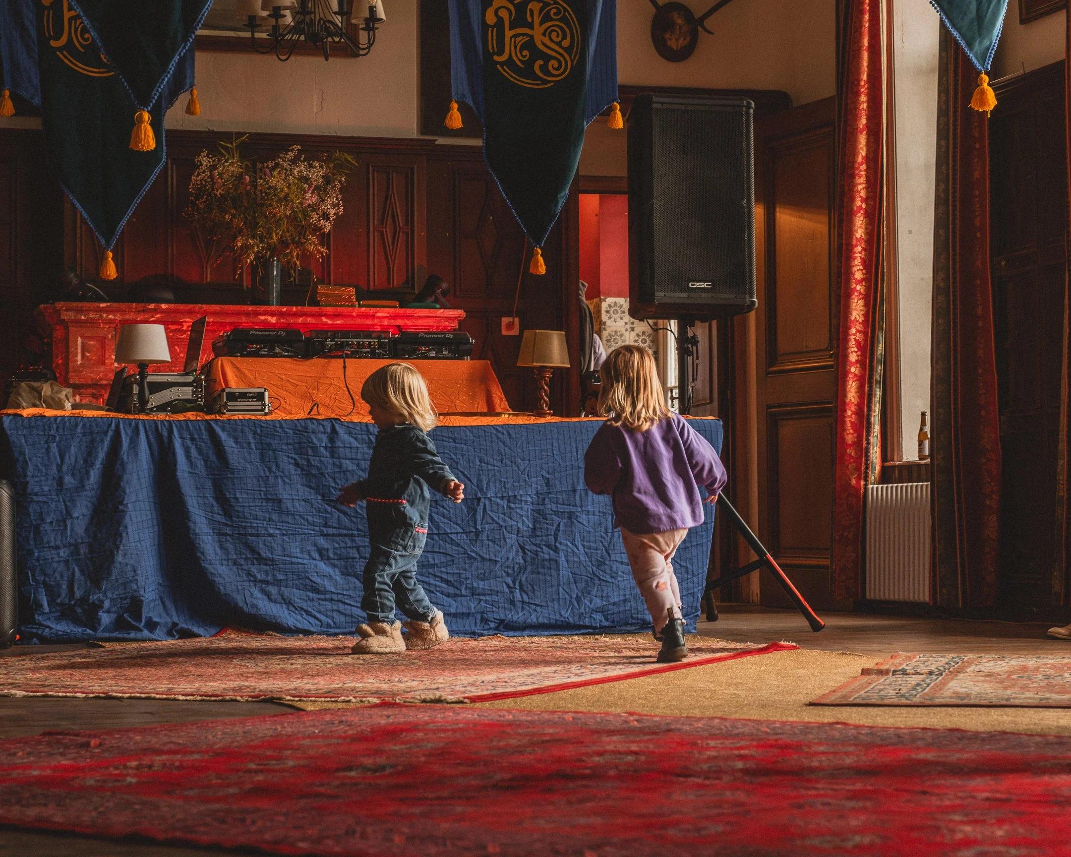Twee jonge kinderen dansen en spelen in een feestzaal met houten wanden, grote ramen met rode gordijnen, en een tafel met DJ-gear en decoraties.