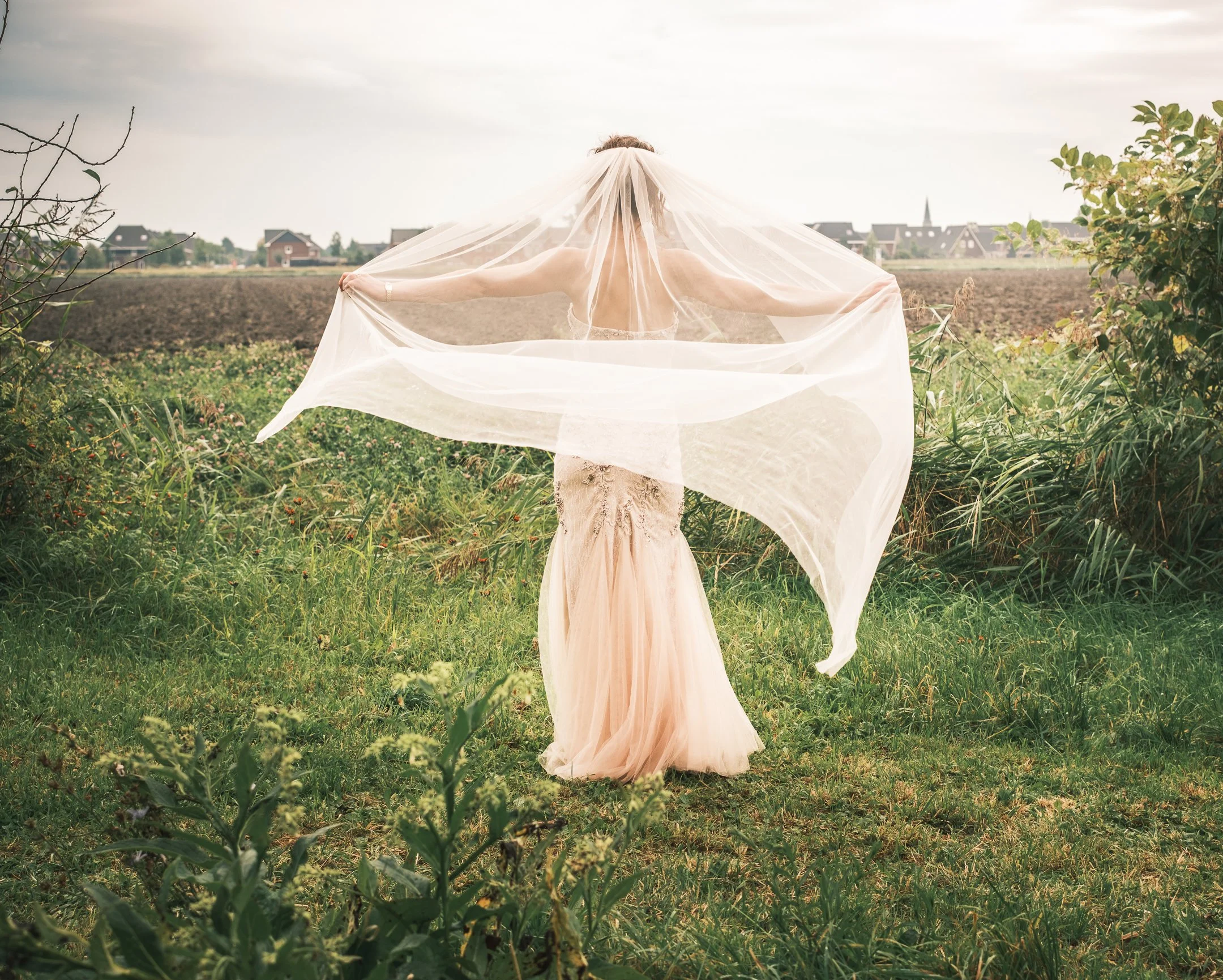 Vrouw in een trouwjurk met sluier staat in een veld, naar de horizon kijkend.