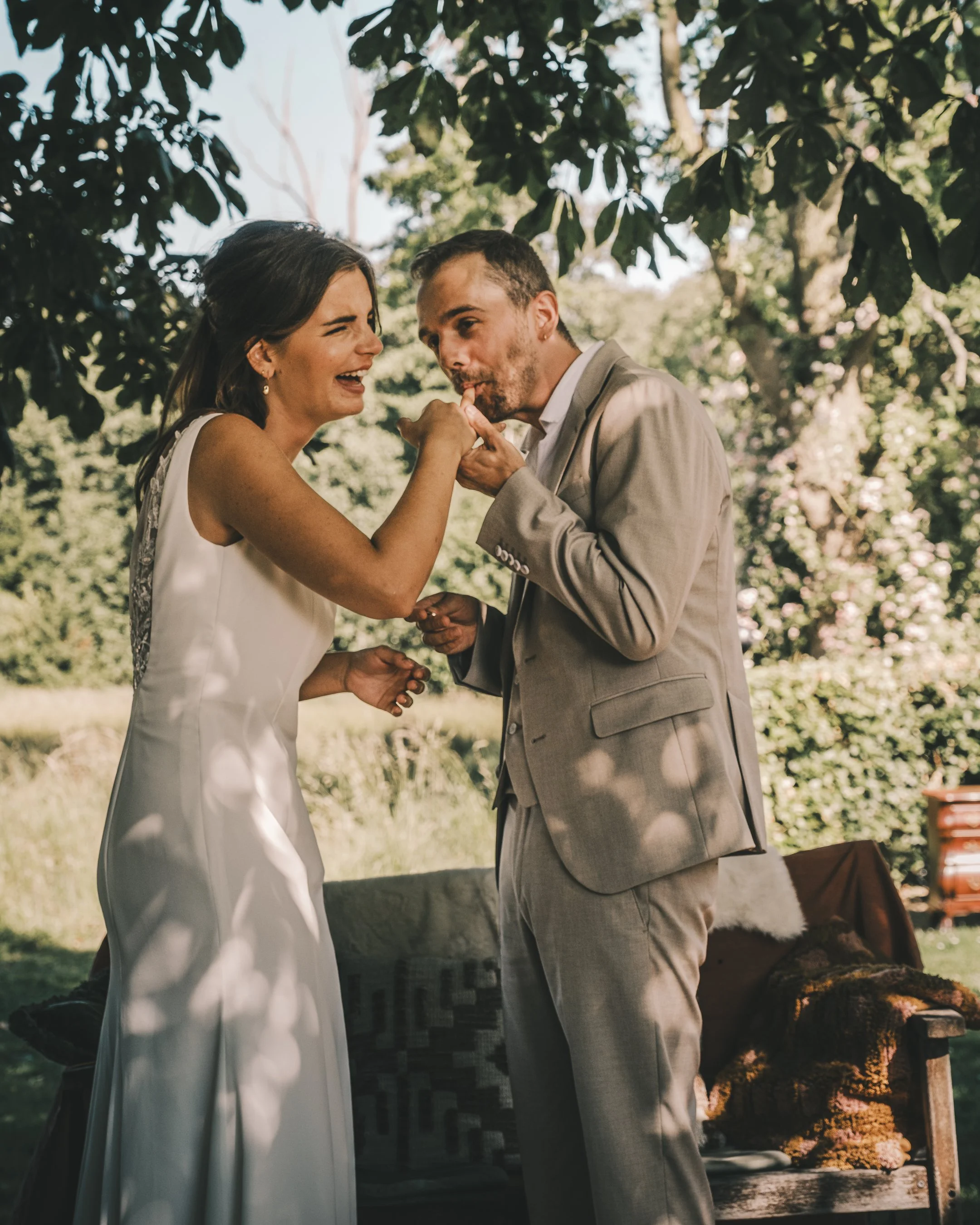 Een bruid en bruidegom tijdens hun huwelijk, de bruidegom kust de hand van de bruid terwijl ze lachen, onder de schaduw van bomen in een tuin.