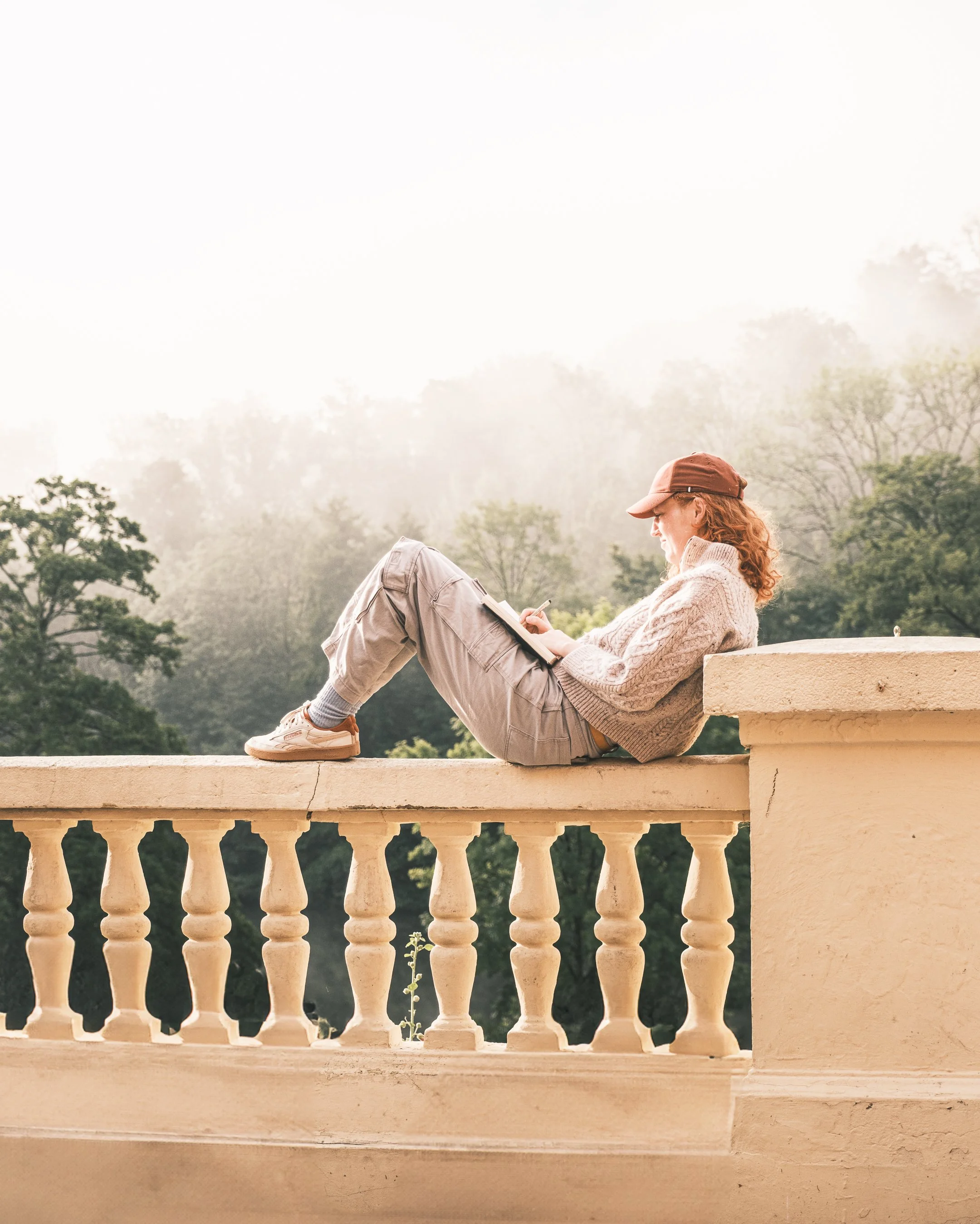 A woman with curly red hair, wearing a beige sweater, khaki pants, a brown cap, and sneakers, sitting on a stone balcony ledge, writing or drawing in a notepad, with a misty green forest in the background.