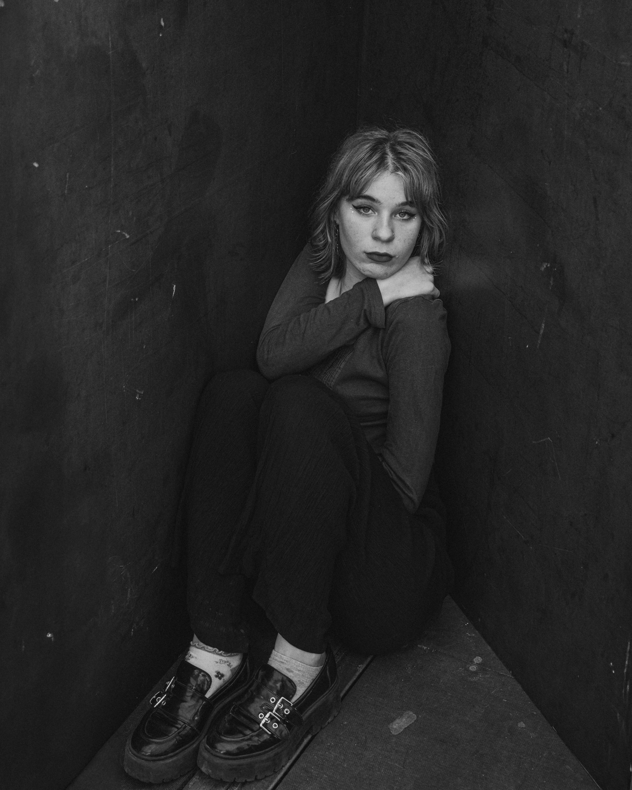 Zwarte en witte foto van een jonge vrouw met licht gekrulld haar, zittend in een hoek, met haar benen tegen haar buik en haar hand op haar schouder, met een serieuze blik.