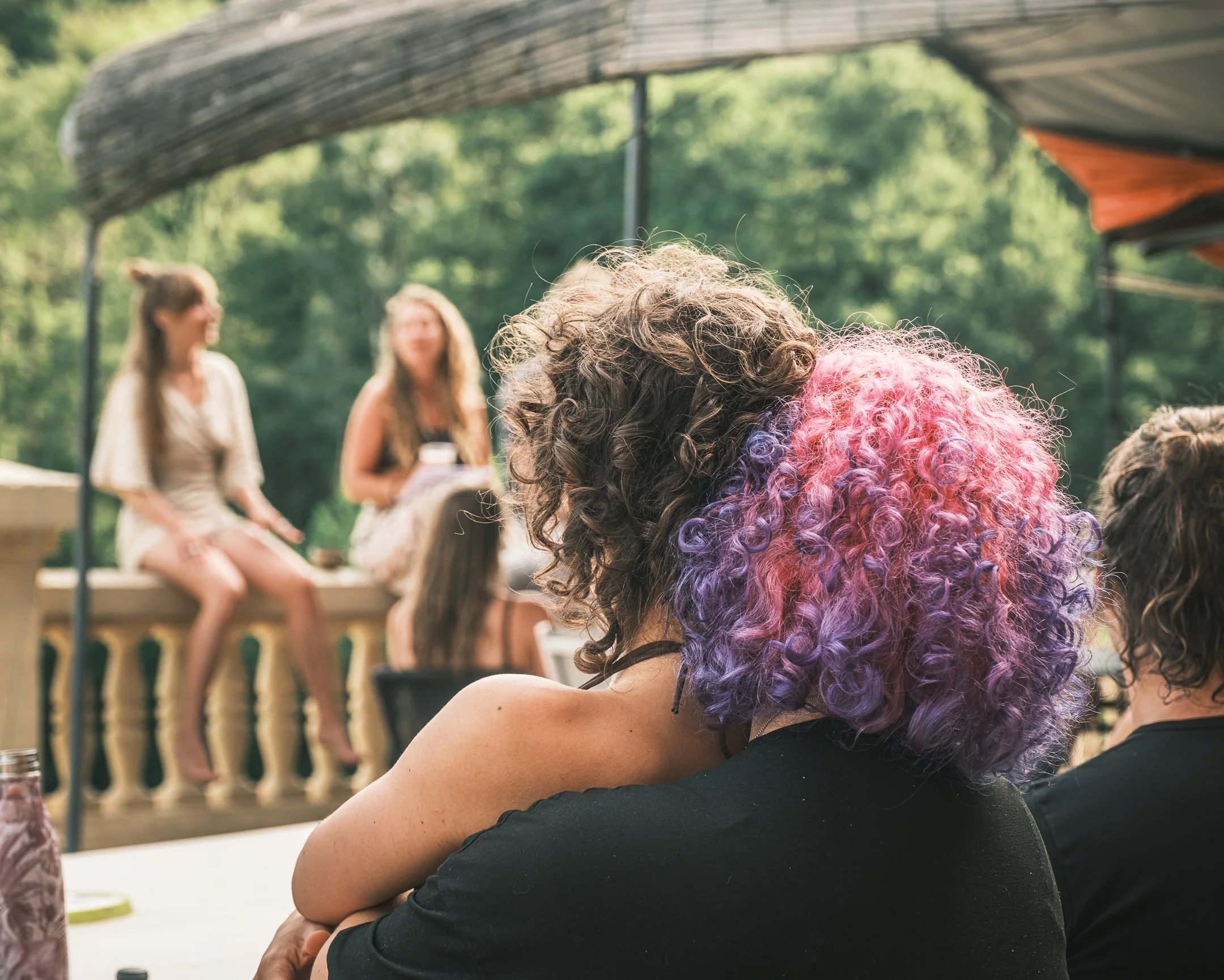 Close-up van een persoon met gekruld haar dat van roze naar paarse kleuren overgaat, zittend en naar een groep mensen die op een podium zitten te kijken, in een groene, bosrijke omgeving.