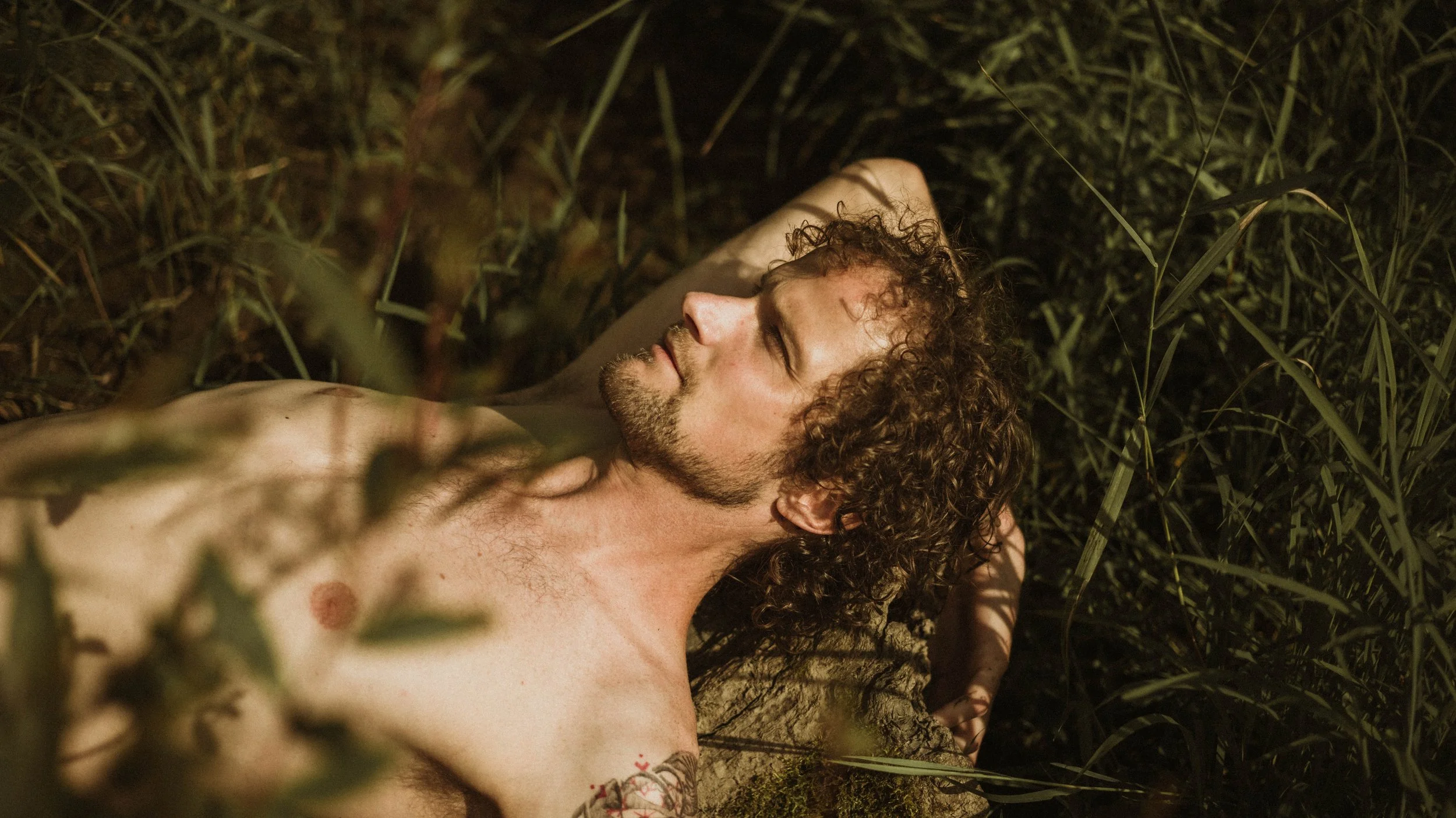 Natuurlijke scène met een naakte man met krullend haar liggend op de grond tussen gras en plantaardig groen, met ogen dicht en een relaxte uitstraling.