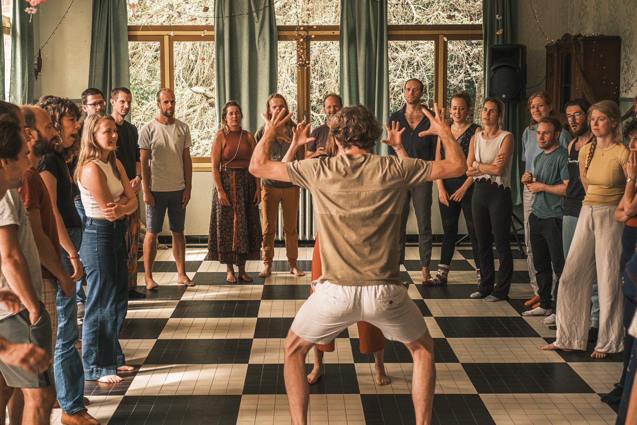 Groep mensen die naar een man luisteren die vooraan staat en met armen en benen beweegt, mogelijk tijdens een workshop of optreden.