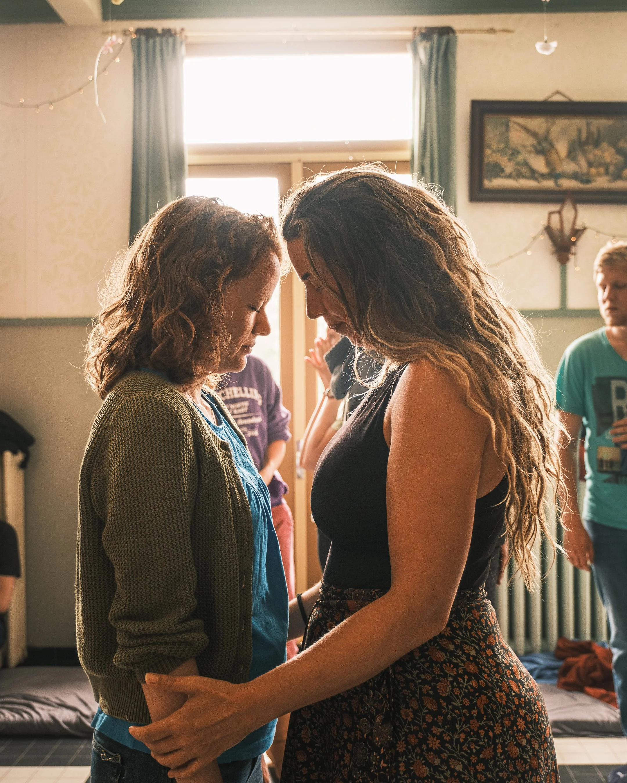 Two women stand closely together with their foreheads touching, holding hands, in a room with sunlight coming through a window behind them. Several other people are in the background.